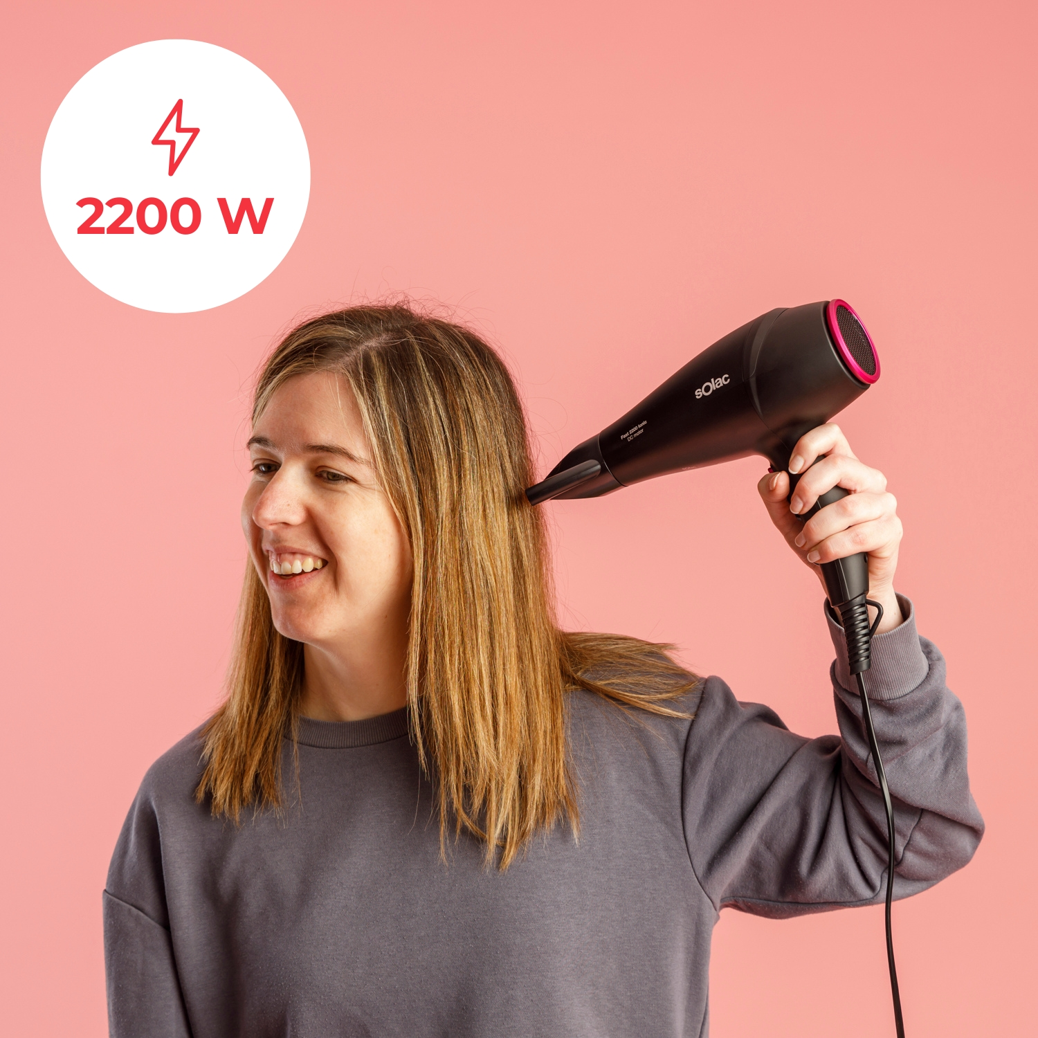 Una mujer se seca el pelo rubio con un secador negro. Se indica una potencia de 2200W.