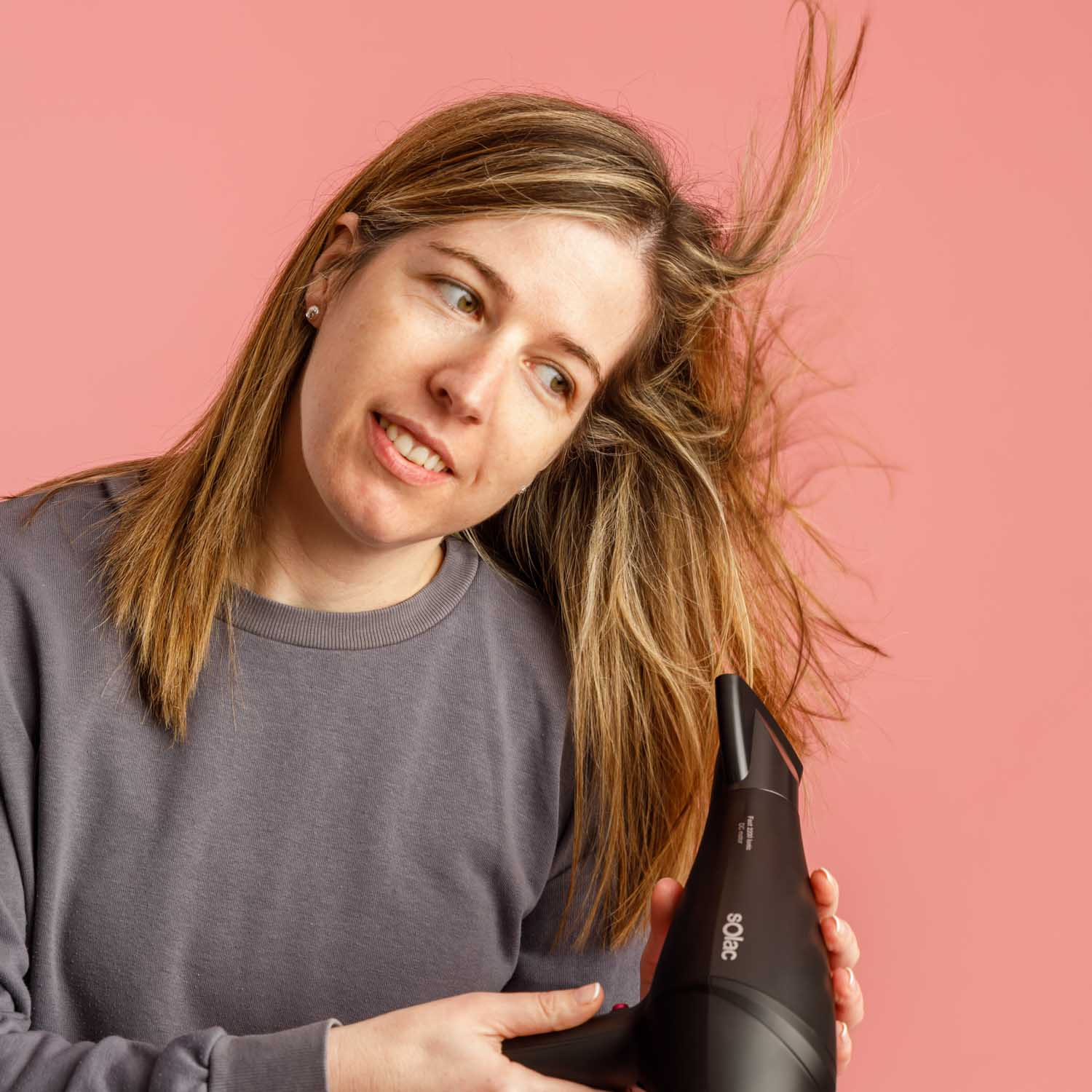 Una mujer se seca el pelo con un secador negro sobre fondo rosa.
