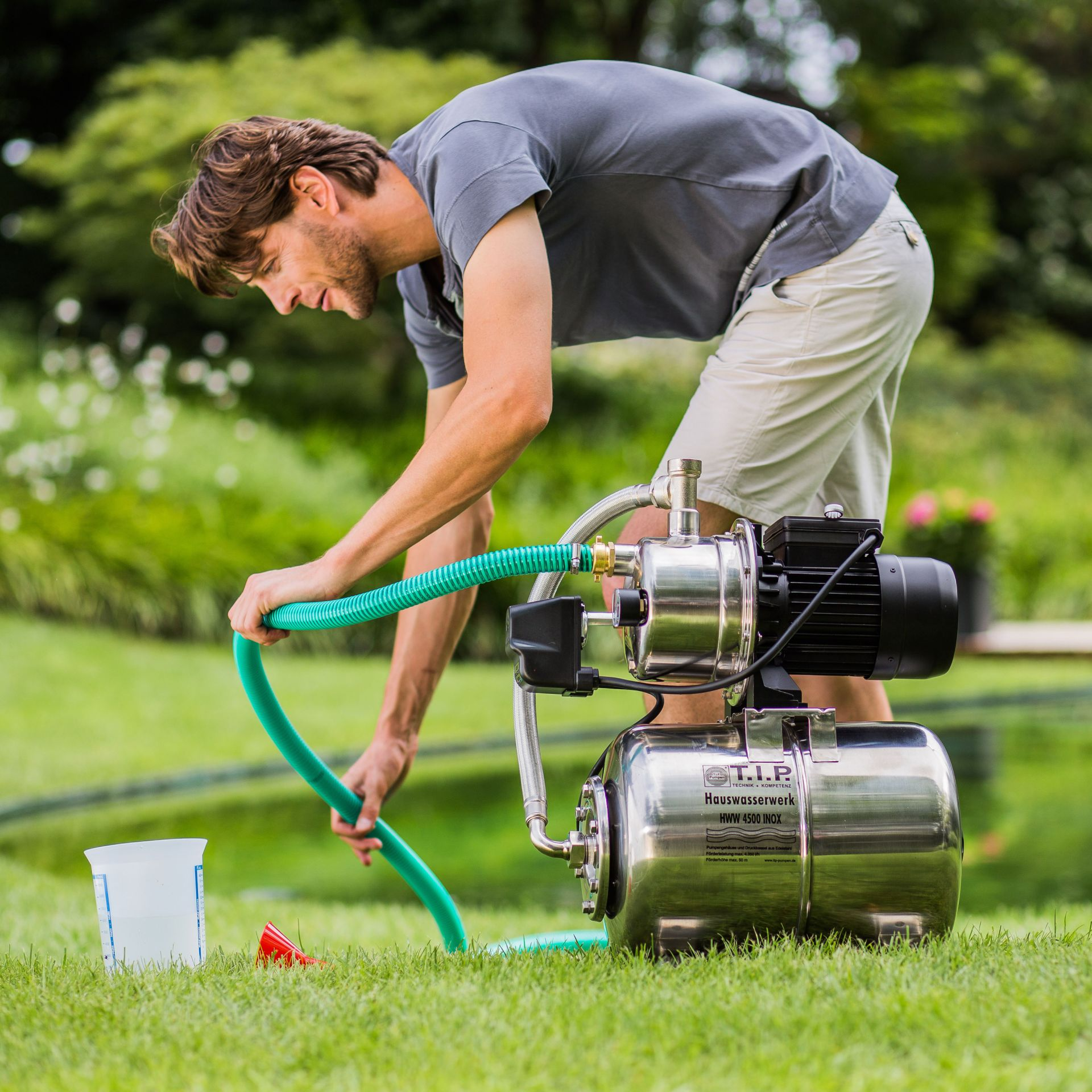 Mann stellt Gartenschlauch an Edelstahl-Hauswasserwerk auf grünem Rasen auf.