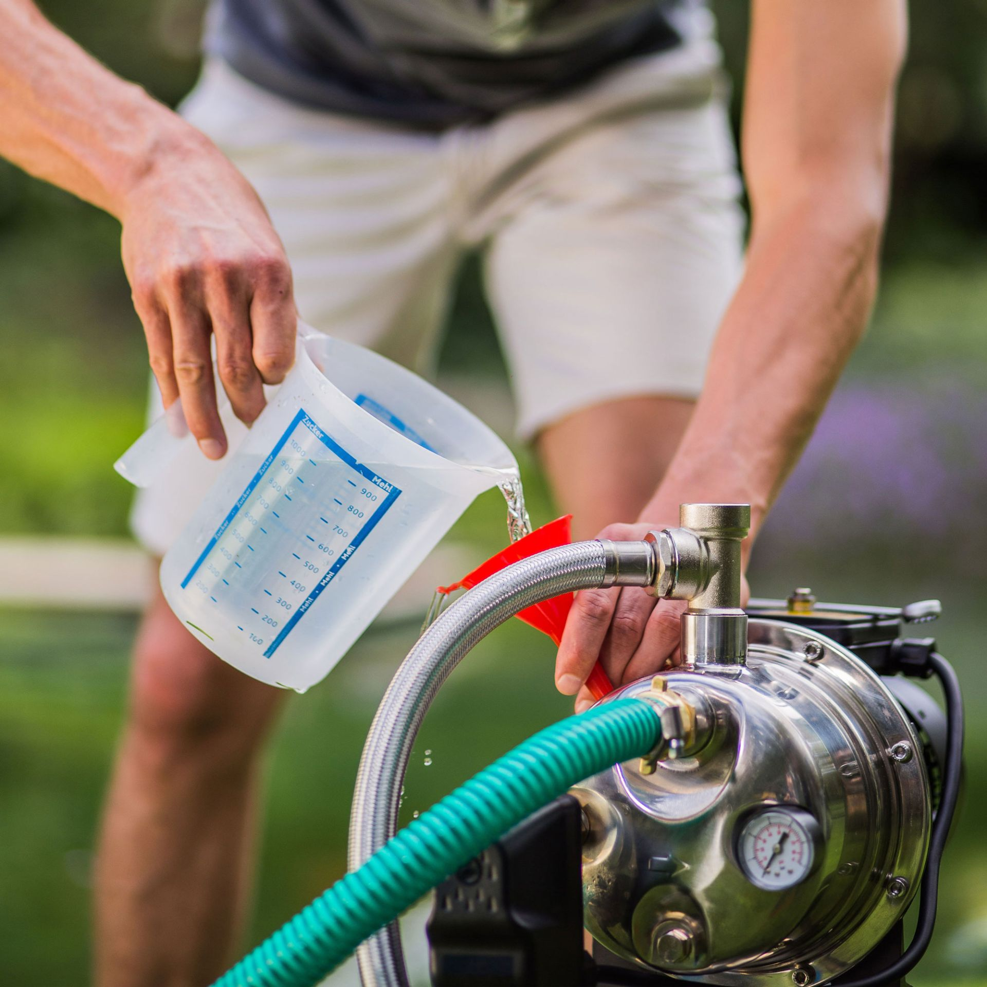Person gießt Wasser aus Messbecher in eine Pumpe mit Schlauch.
