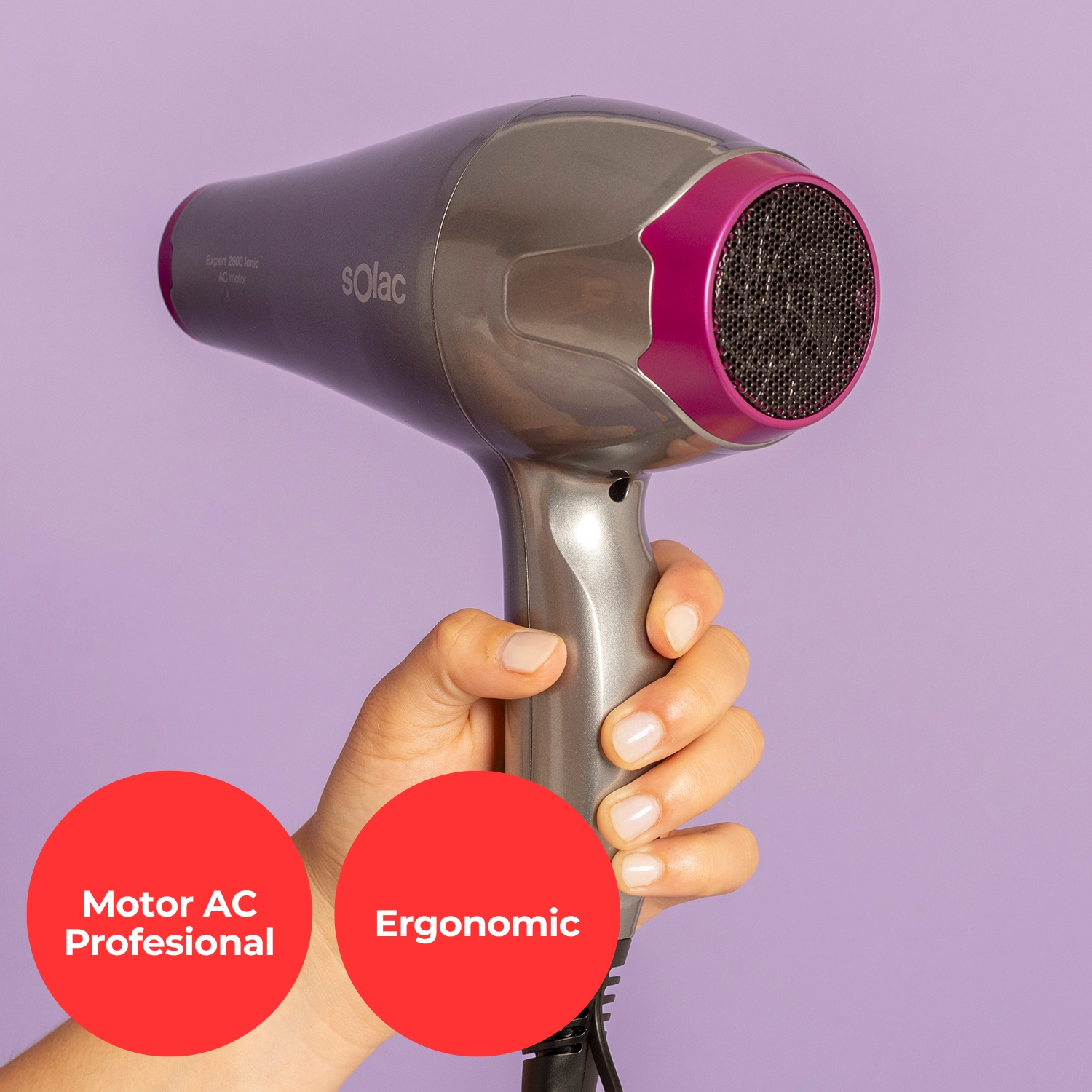 Una mano sostiene un secador de pelo gris con acento rosa sobre fondo morado.