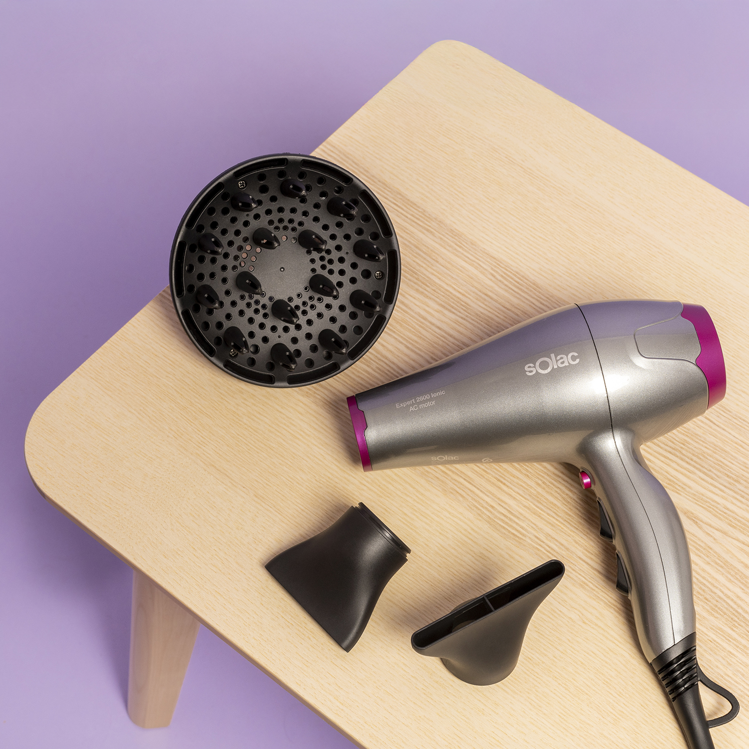 Un secador de pelo gris con detalles rosas, difusor y dos boquillas en mesa madera.