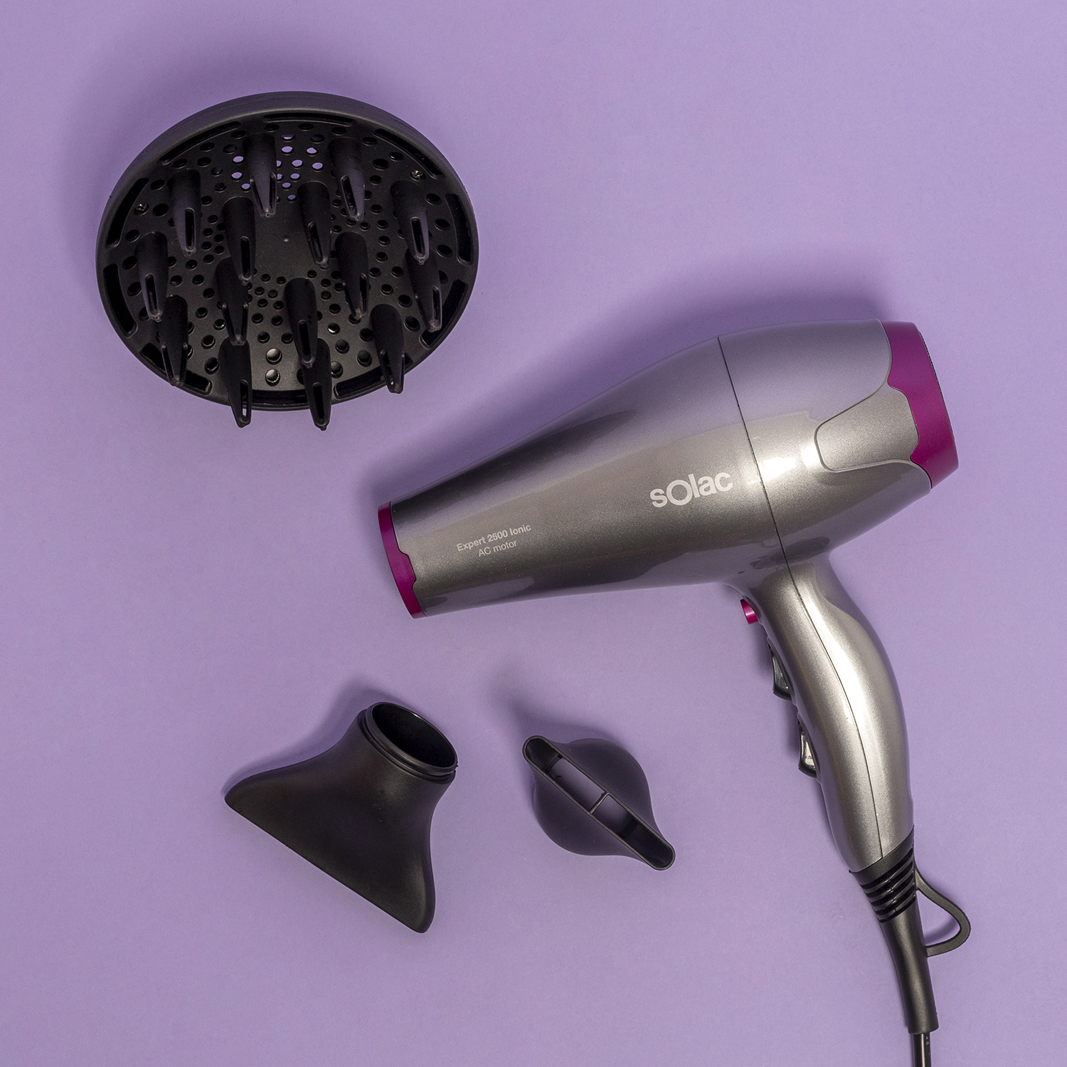 Un secador de pelo gris con detalles rosas, difusor y dos boquillas sobre fondo morado.