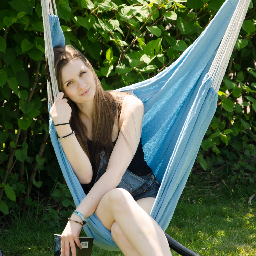 Eine Frau sitzt in einer blauen Hängematte im Freien vor grünen Büschen.