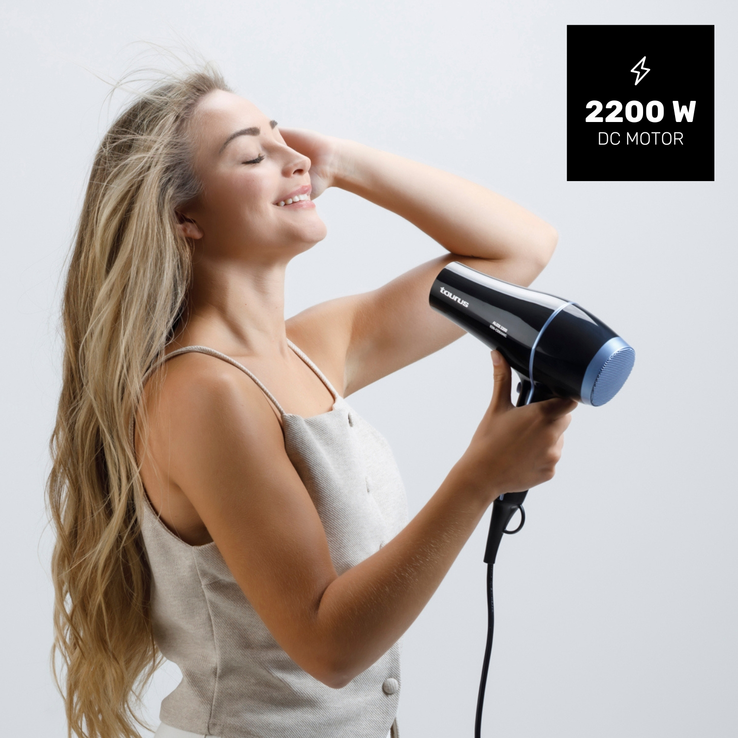 Mujer con pelo rubio largo se seca el pelo con un secador negro. Motor DC de 2200 W.