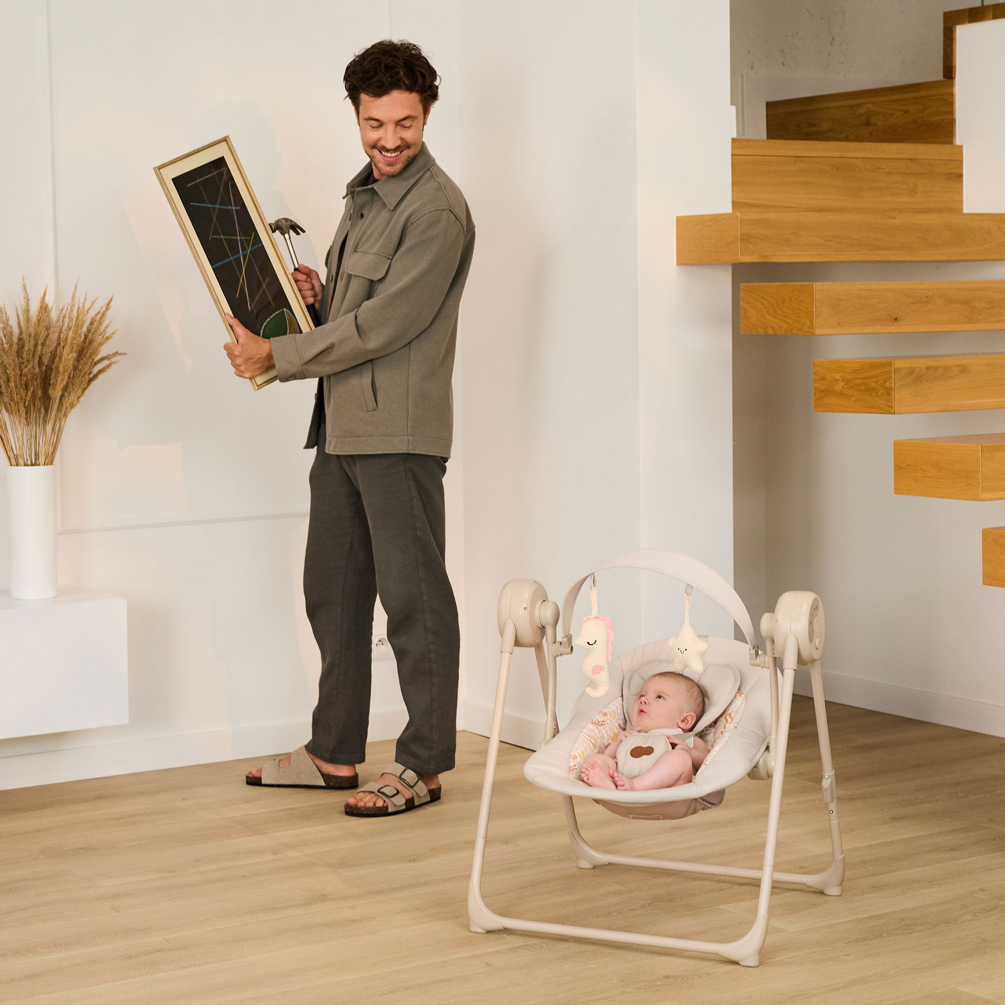 Mann hält Bild und Hammer; Baby schaukelt in einem Babywippen. Holztreppe im Hintergrund.