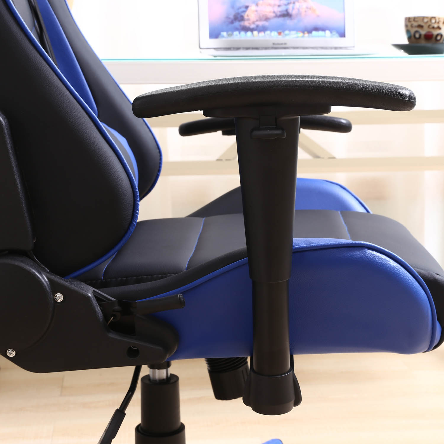 Una silla gamer negra y azul con reposabrazos en primer plano. Un portátil y una taza al fondo.