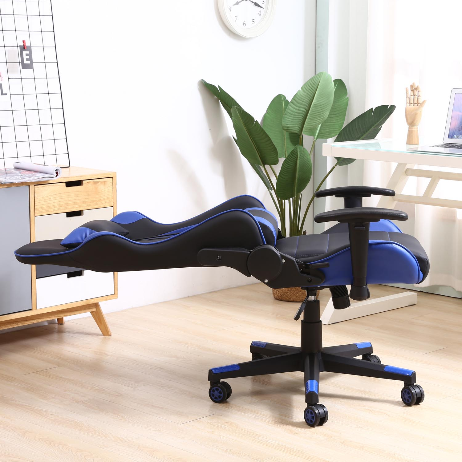 Una silla gamer negra y azul con reposapiés en una habitación con escritorio y planta.