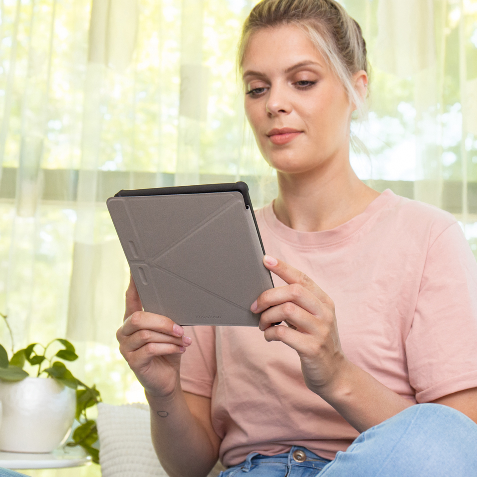 Femme en t-shirt rose et jean tient une tablette grise avec support plié.