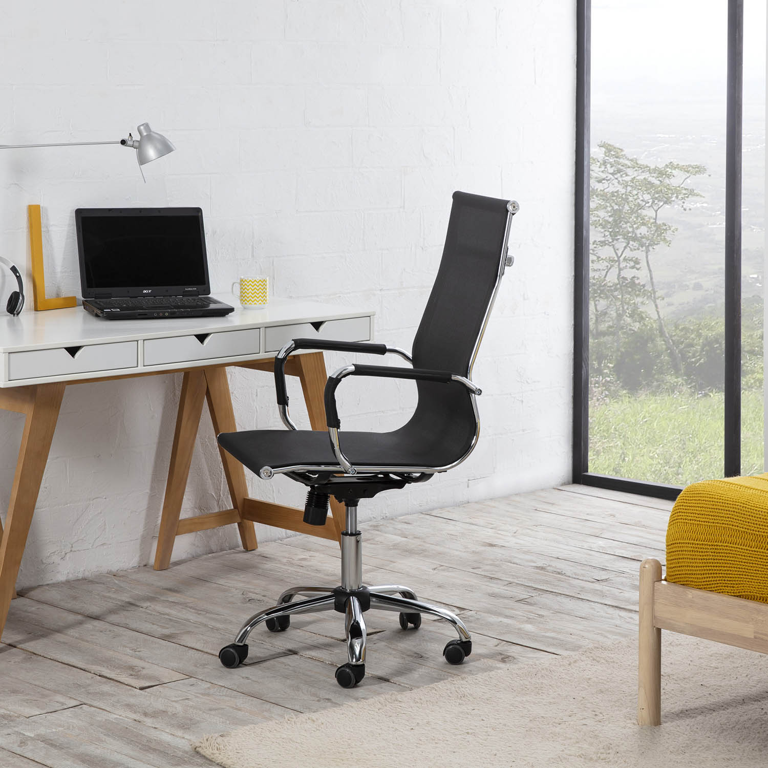 Una silla de oficina negra junto a un escritorio blanco con portátil y taza amarilla.