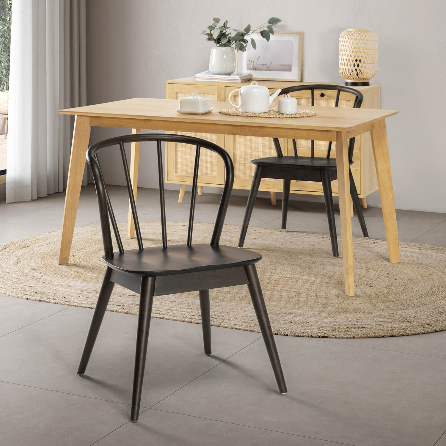 Una mesa de comedor de madera clara con sillas oscuras sobre una alfombra de yute. Un aparador con decoración detrás.