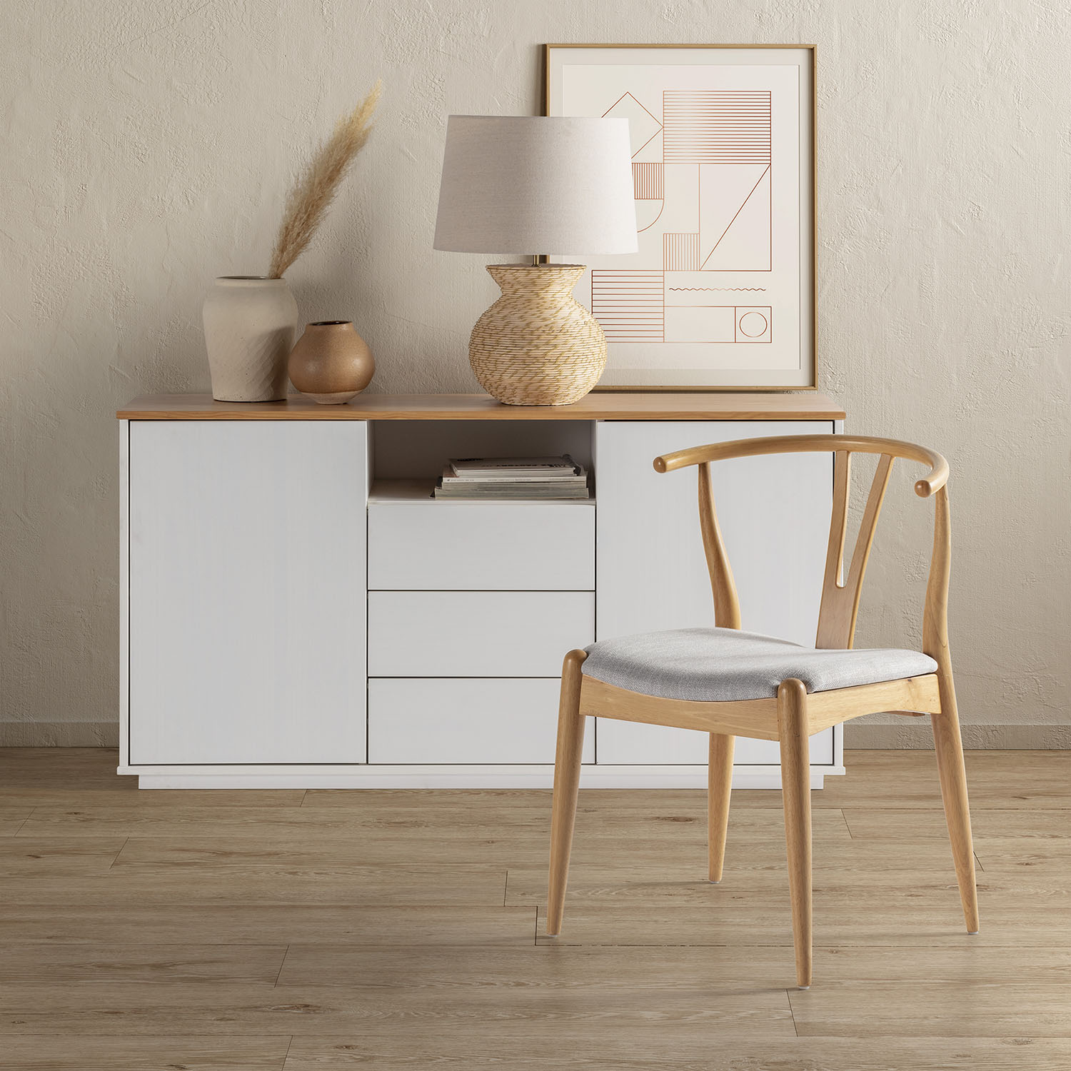 Un aparador blanco con decoración, lámpara y arte abstracto enmarcado, con una silla de madera delante.