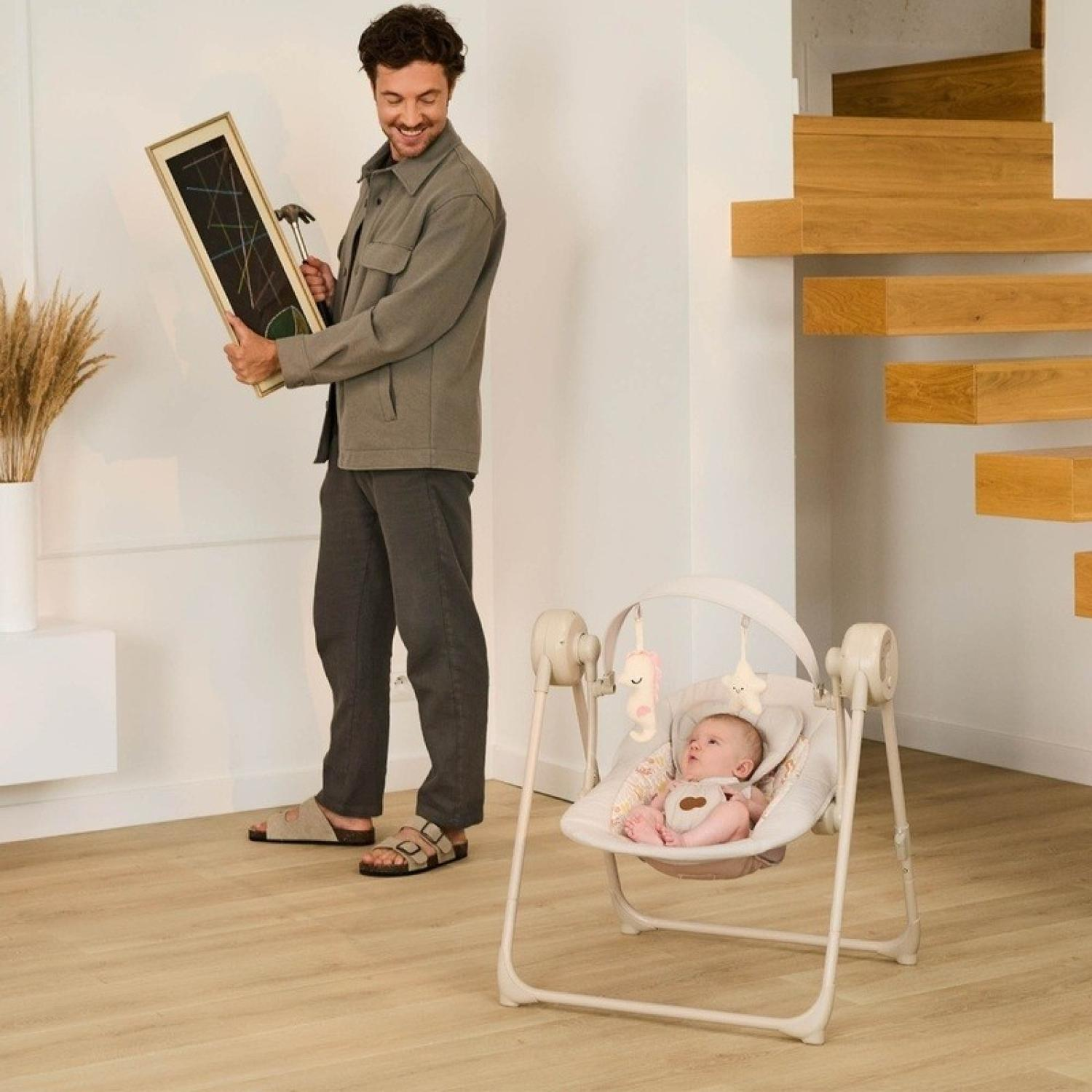 Mann hält gerahmte Kunst und Hammer neben einer Babyschaukel mit Baby darin.