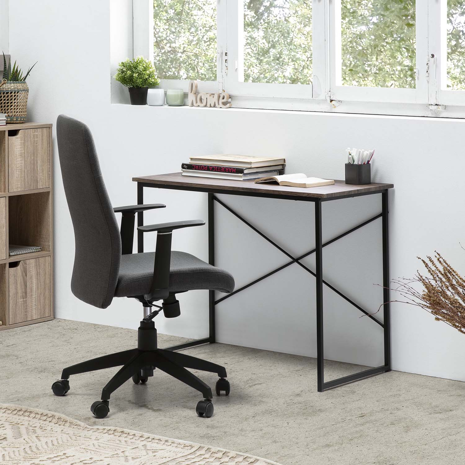 Una silla de oficina gris oscuro junto a un escritorio de madera con libros y bolígrafos cerca de una ventana.