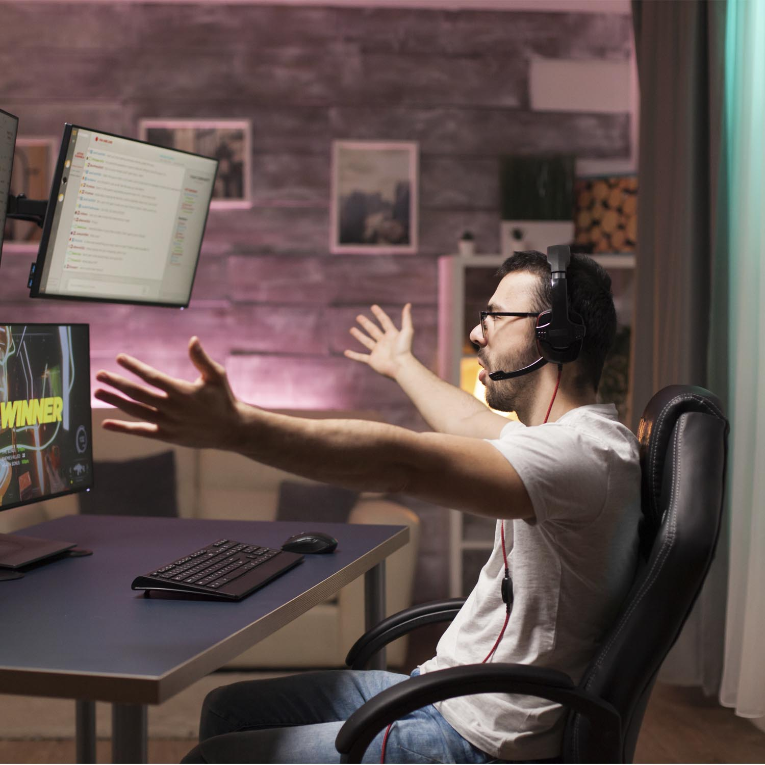 Un hombre con auriculares se sienta en un escritorio con dos monitores, gesticulando.