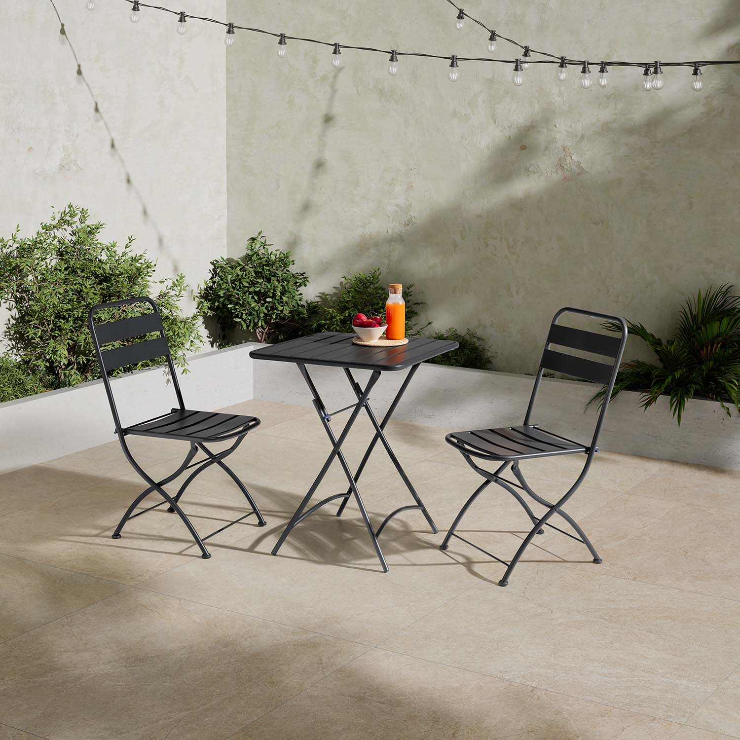 Una mesa plegable negra con dos sillas al aire libre, iluminada por luces de cadena.