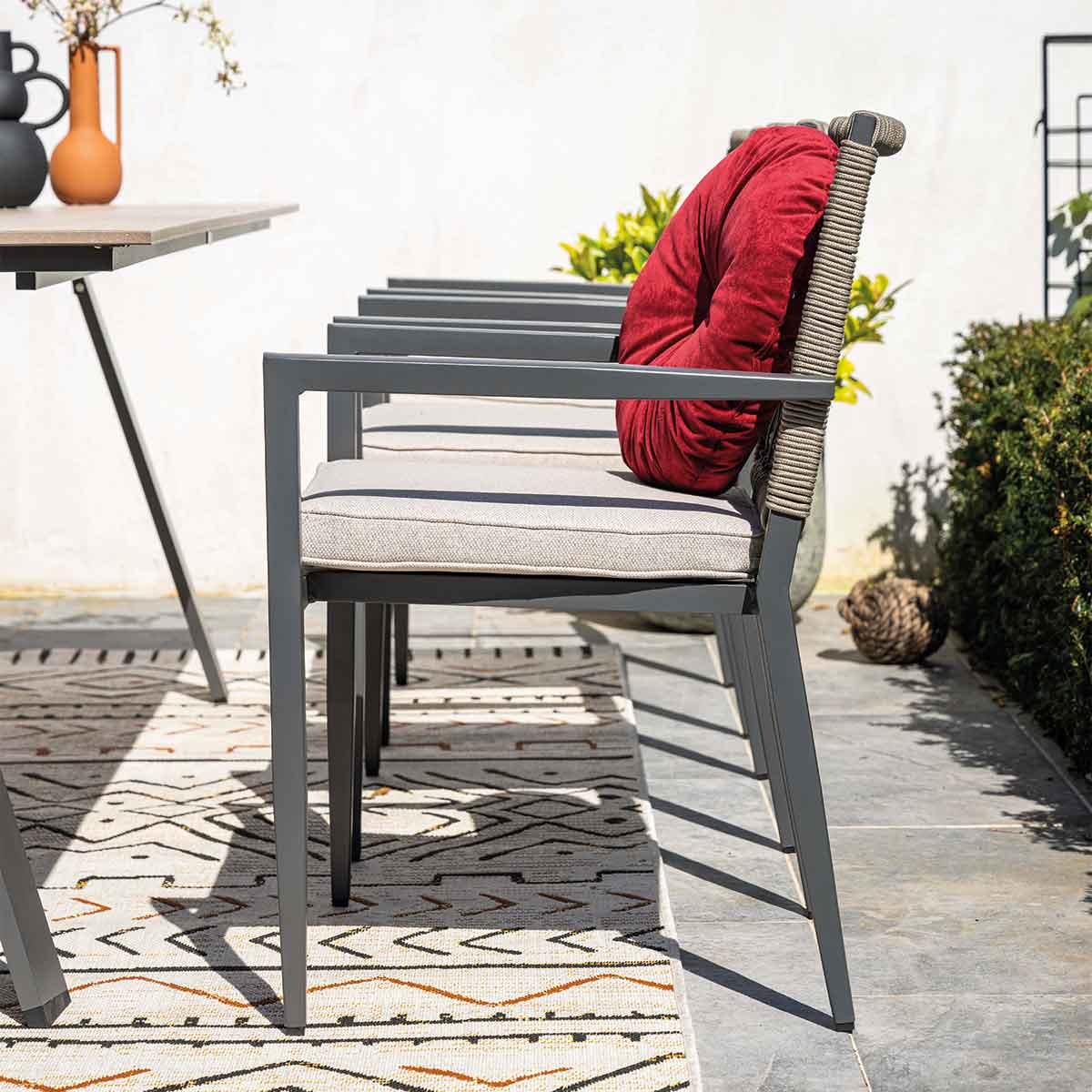 Grijze buitenstoelen met lichte kussens en een rood rugkussen op een vloerkleed.