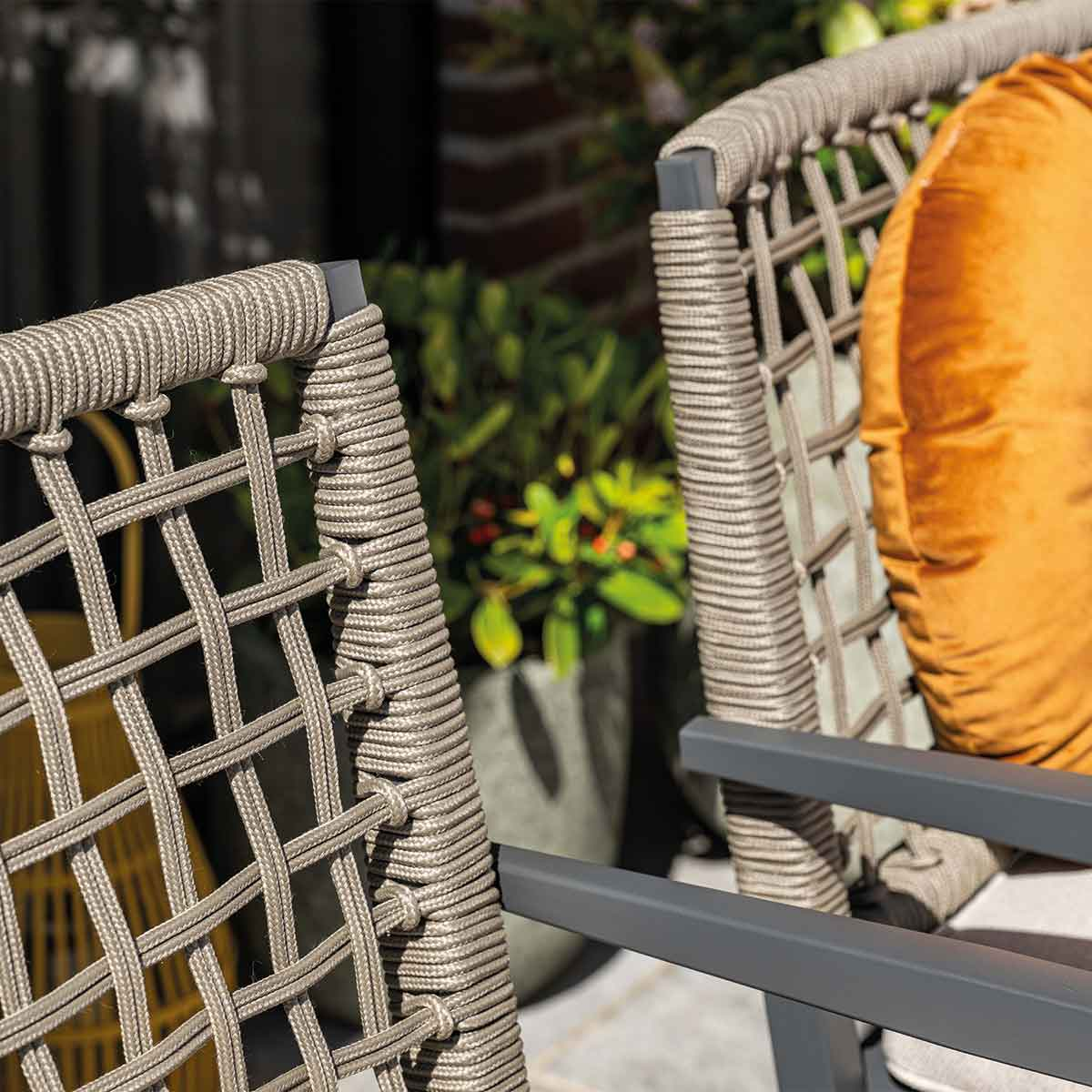 Twee buitenstoelen met gevlochten touwrug, één met oranje kussen. Groene planten op achtergrond.