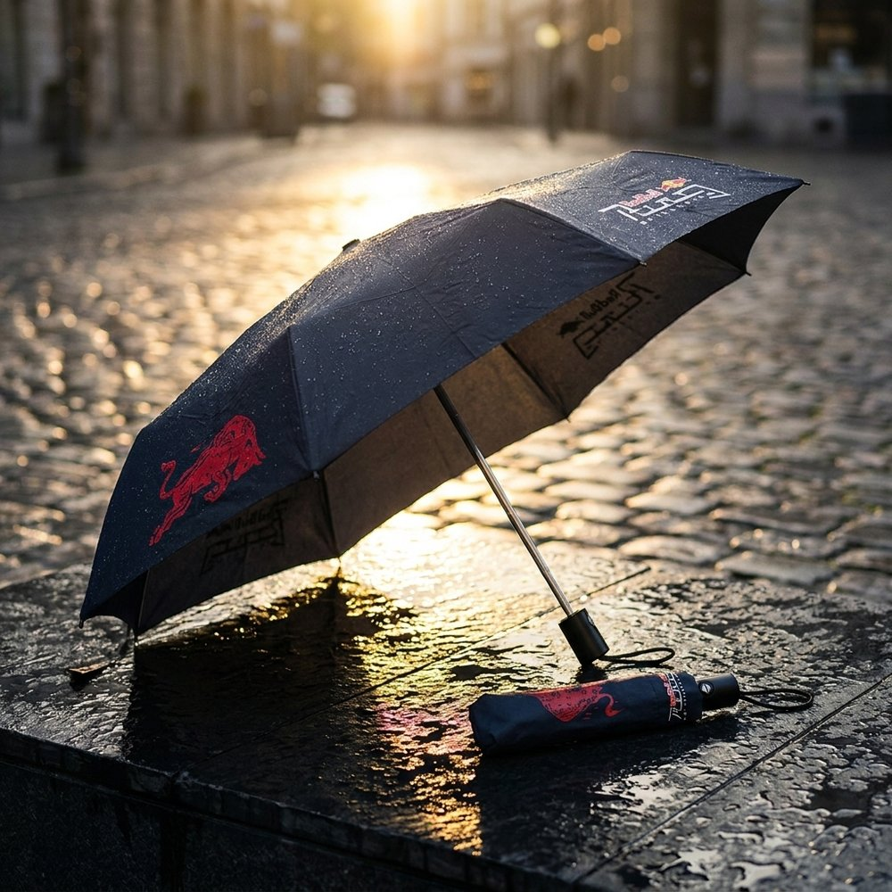 Un ombrello nero bagnato con logo Red Bull su una strada acciottolata bagnata al tramonto.