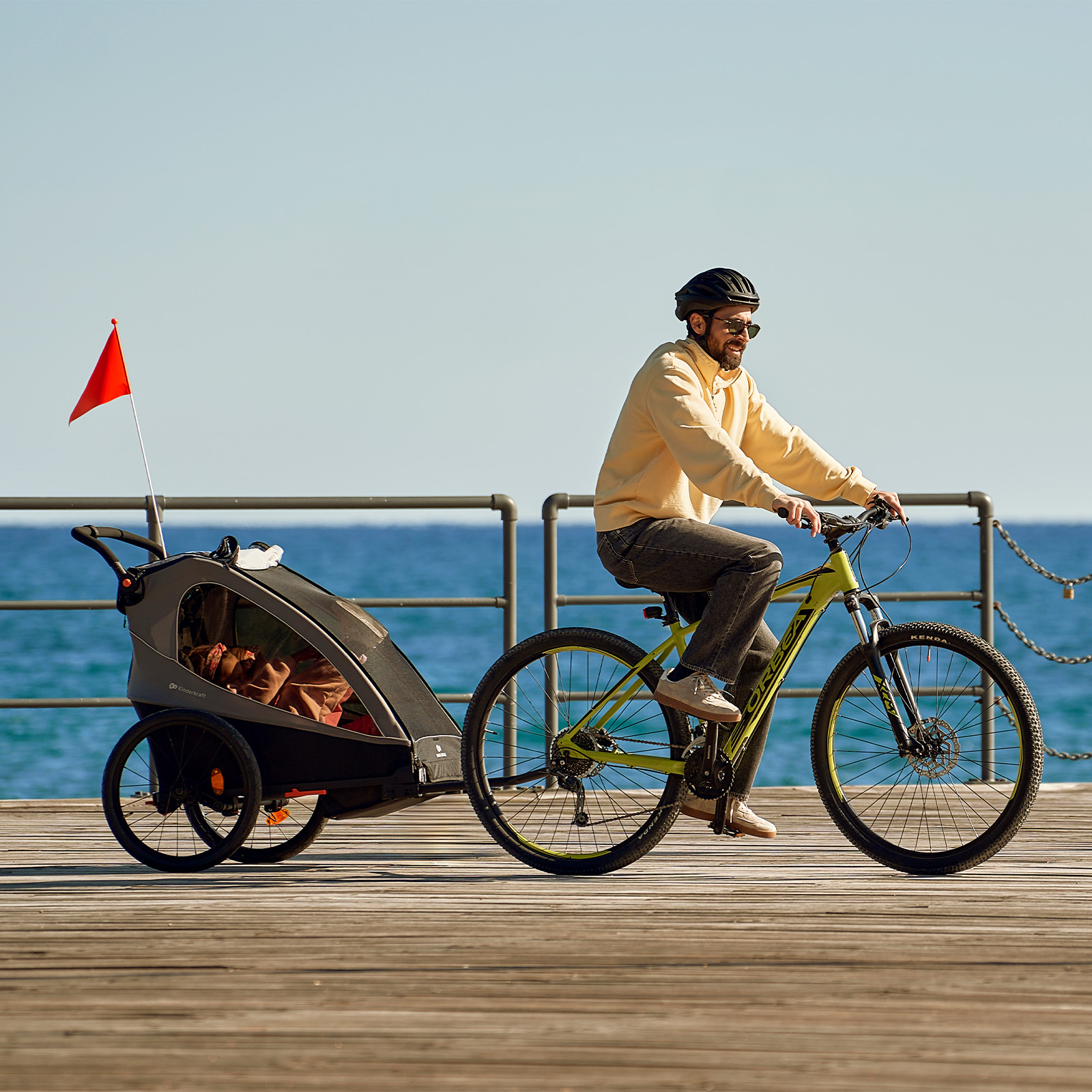 Mann fährt Fahrrad mit Kinderanhänger am Meer entlang auf Holzsteg.