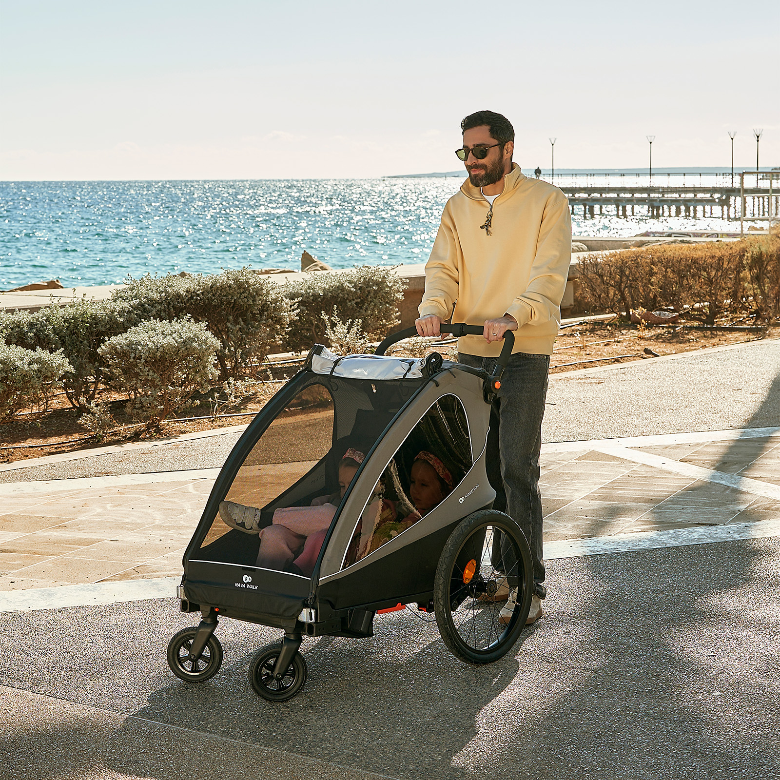 Mann schiebt Kinderwagen mit zwei Kindern am Meer entlang, mit Pier im Hintergrund.