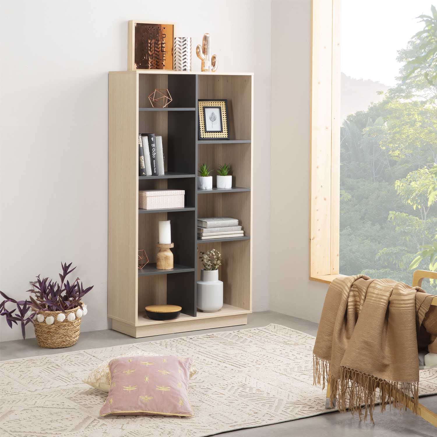 Una estantería con decoración, plantas y libros junto a una ventana con vistas a los árboles.