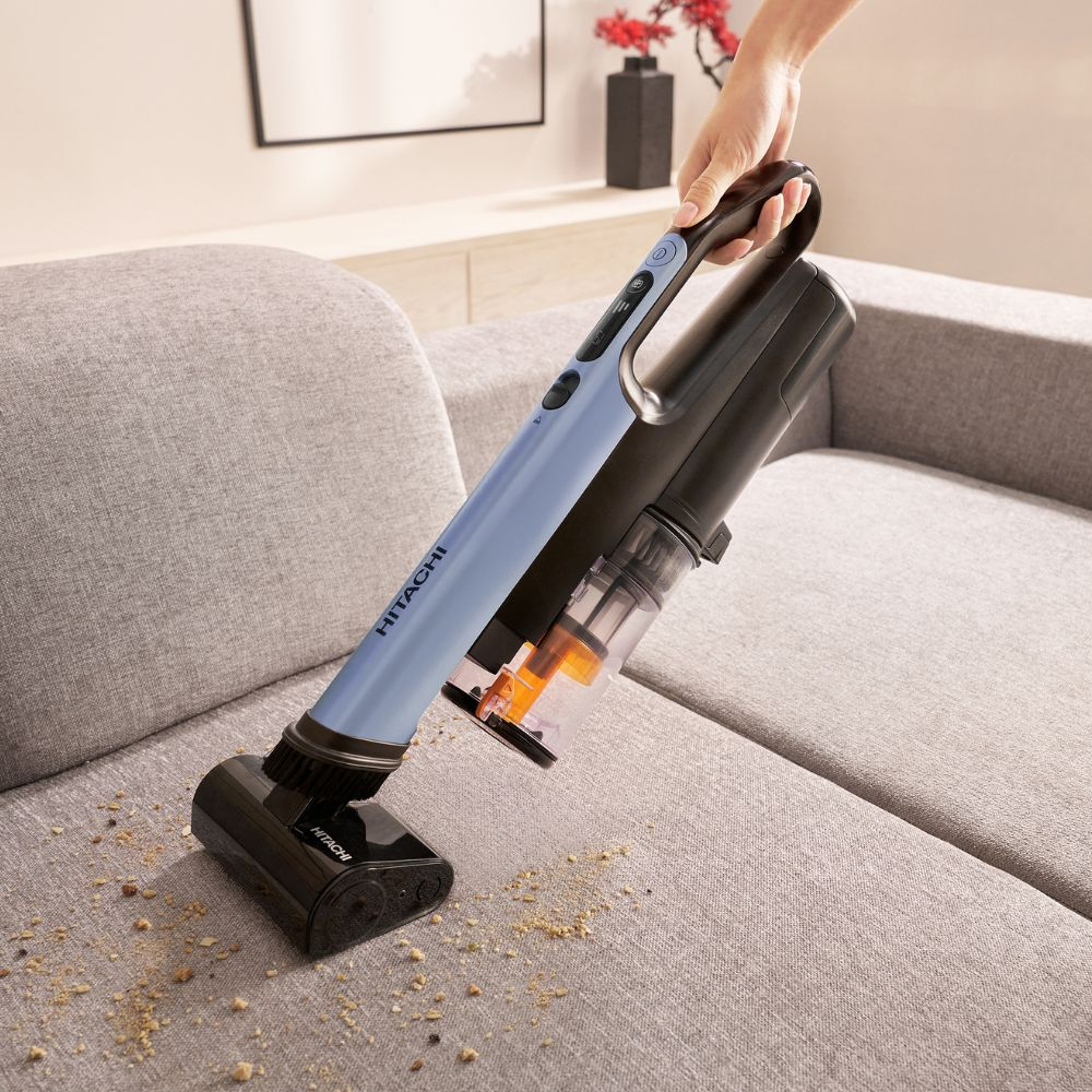 Eine Hand hält einen blauen Hitachi-Staubsauger, der Krümel von einem grauen Sofa saugt.