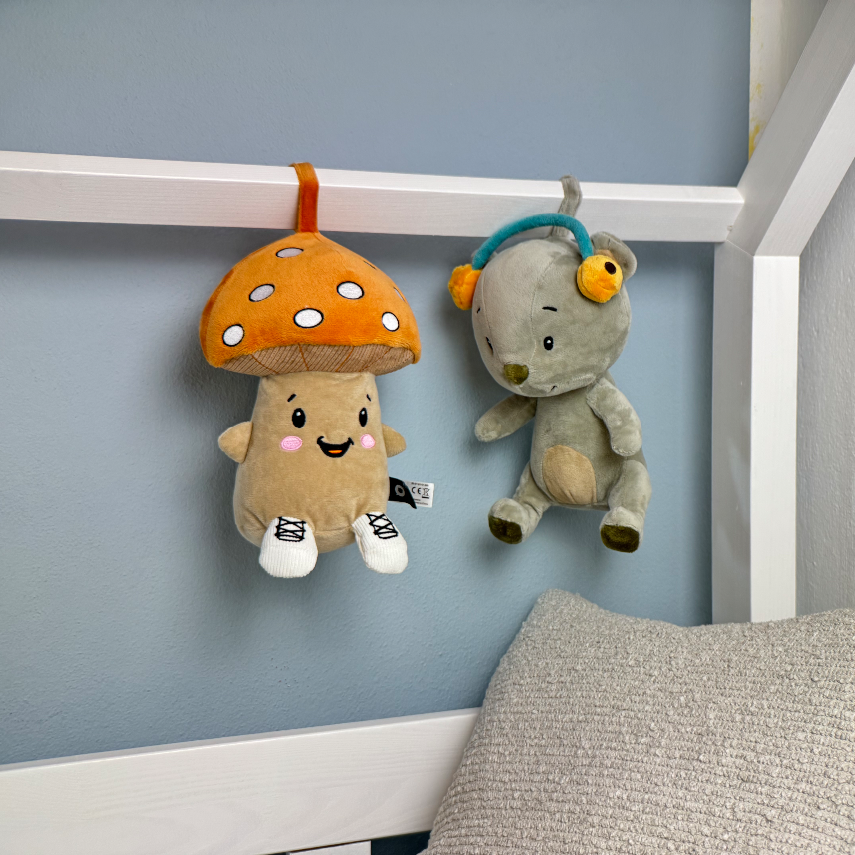 Ein braunes Pilz-Stofftier mit orangenem Hut und ein graues Teddybär-Stofftier mit Kopfhörern hängen an einem weißen Bettrahmen.