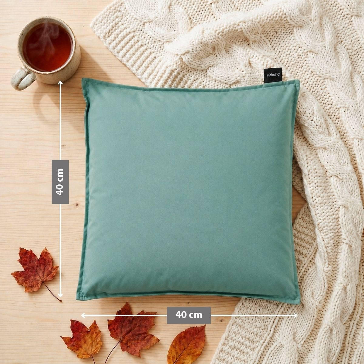 A teal cushion rests on a wooden table beside a cup of tea and autumn leaves.