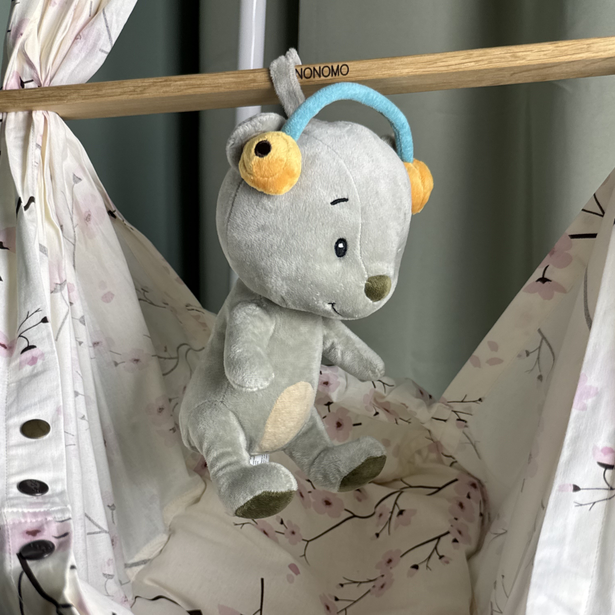 Ein graues Plüschtier mit Kopfhörern hängt über einem Bettchen mit Blumenmuster.