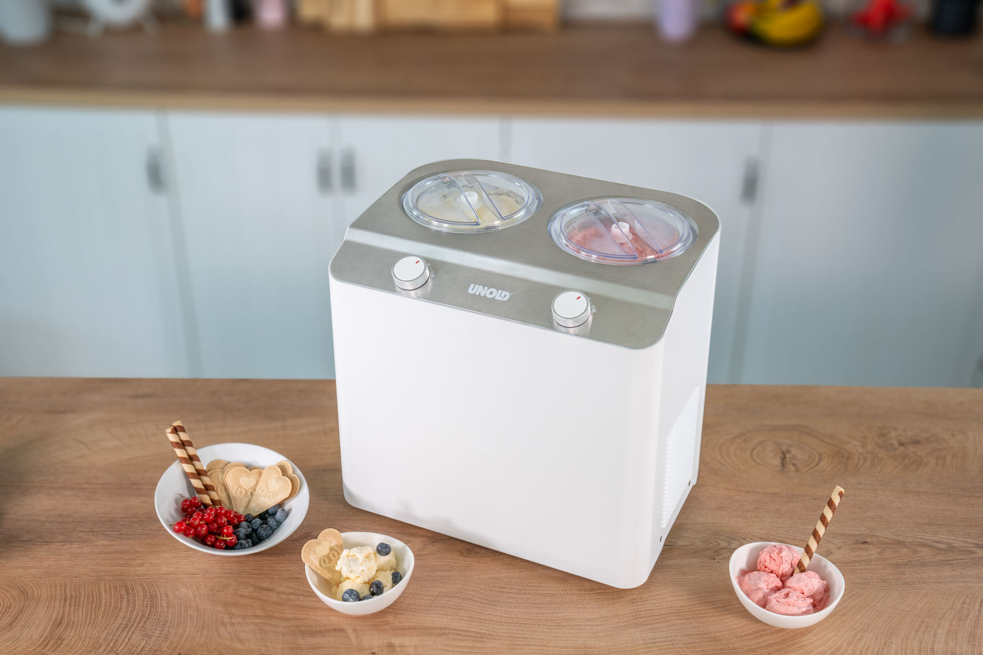 Weiße Eismaschine mit zwei Behältern, daneben Schalen mit Eis, Beeren und Keksen.