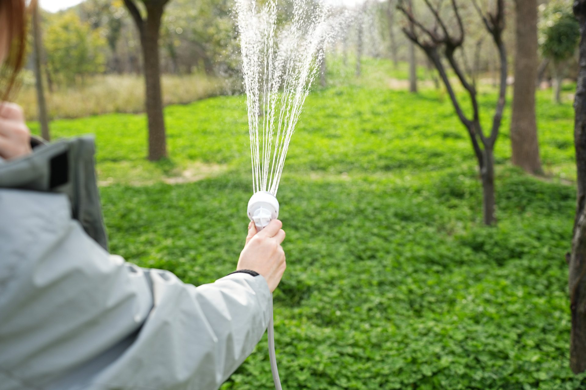 Eine Hand hält ein weißes Duschventil, aus dem Wasser spritzt, auf grünem Gras.