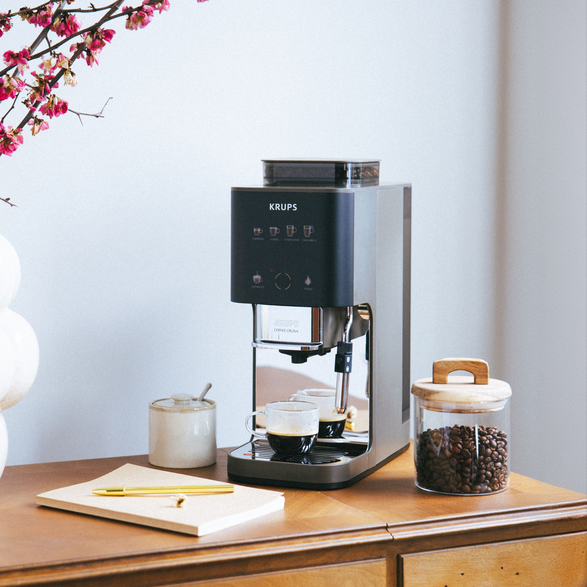 Eine Krups Kaffeemaschine mit zwei Tassen Kaffee, Bohnen und Notizbuch auf einem Holztisch.