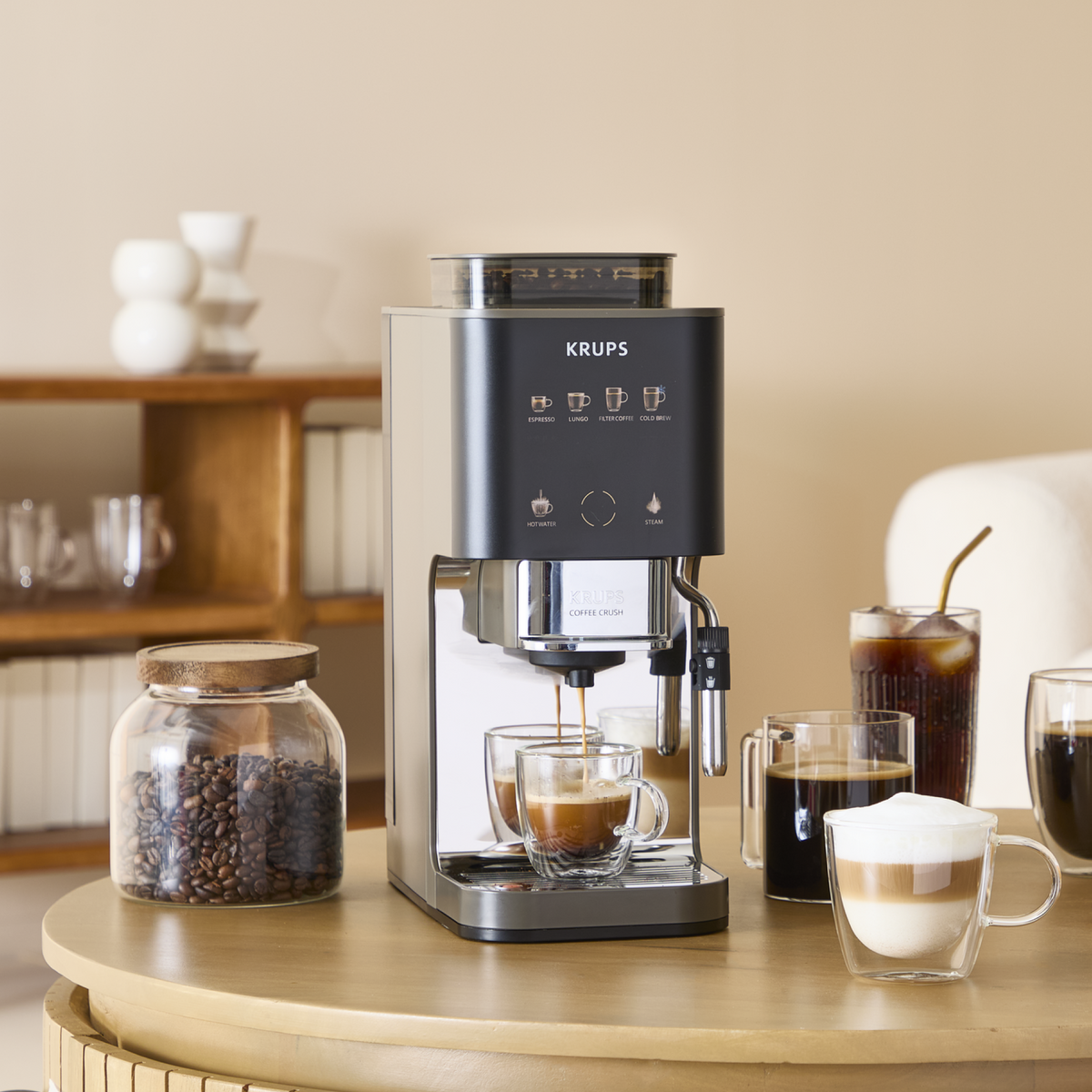 Eine Krups-Kaffeemaschine brüht Kaffee in eine Glastasse, daneben Kaffeebohnen und Getränke.