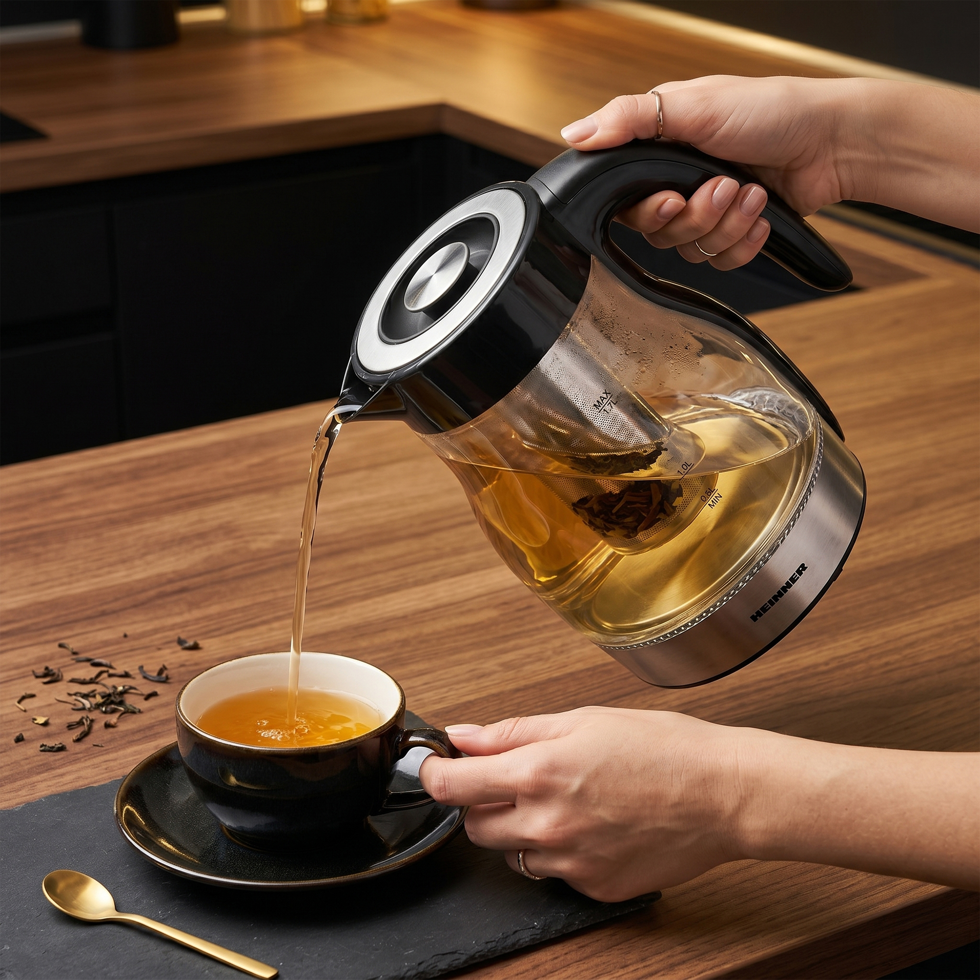 Een hand schenkt thee uit een glazen theepot in een zwarte kop op een houten tafel.