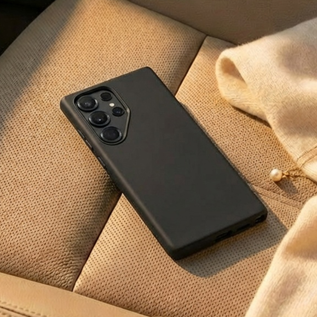 Une coque de téléphone noire repose sur une surface beige texturée, avec un tissu crème plié et un détail perle.