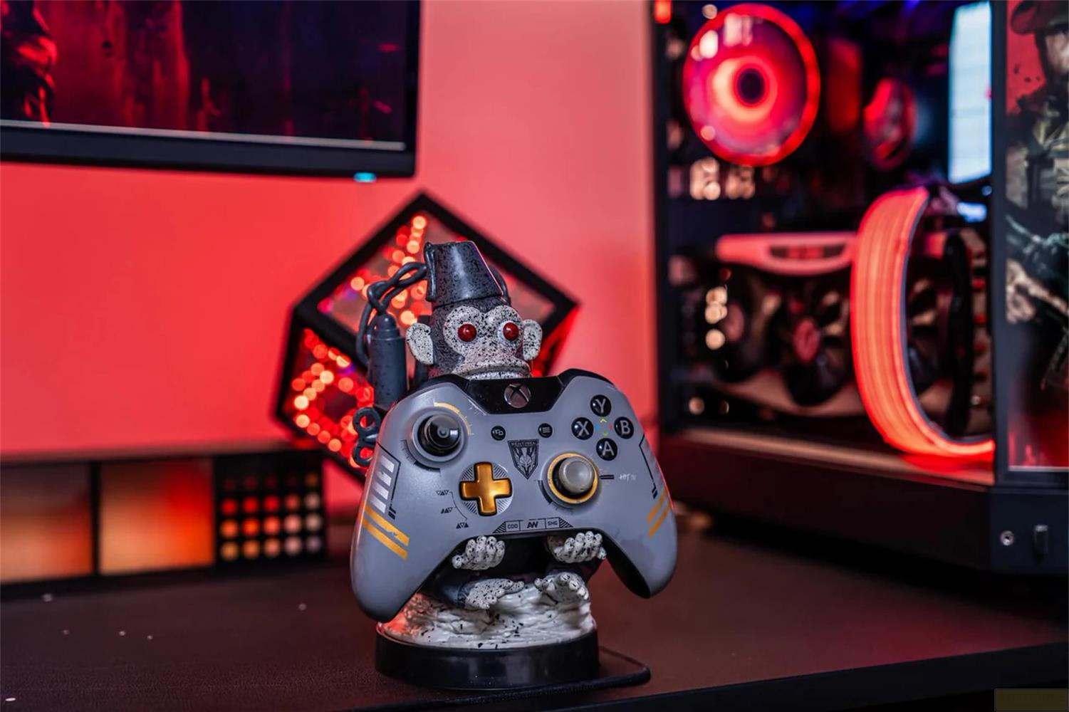 Un mando gris de Xbox lo sostiene una figura de mono, con un PC y un monitor al fondo.