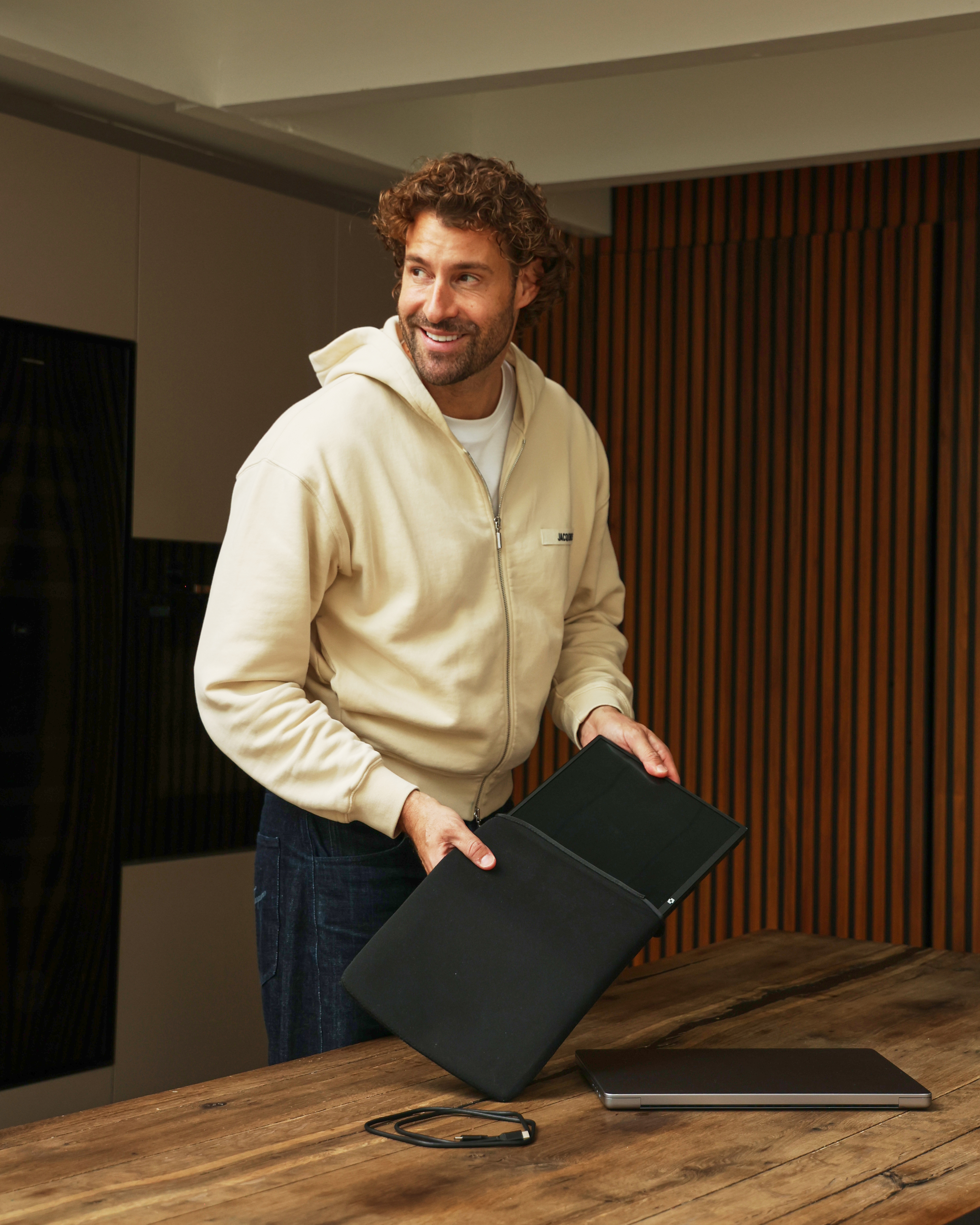 Een man in een beige hoodie en spijkerbroek houdt een zwarte laptoptas vast.