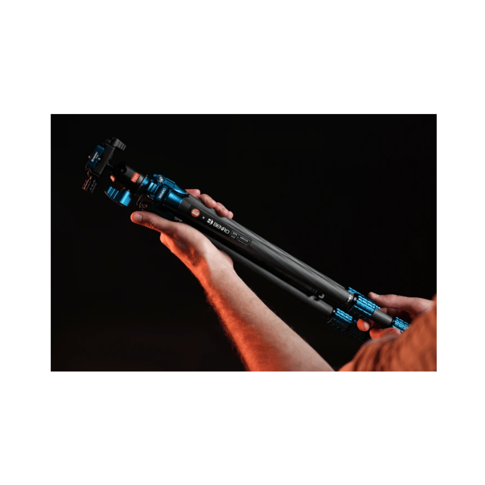 Een persoon houdt een zwart en blauw carbon fiber statief vast tegen een zwarte achtergrond.