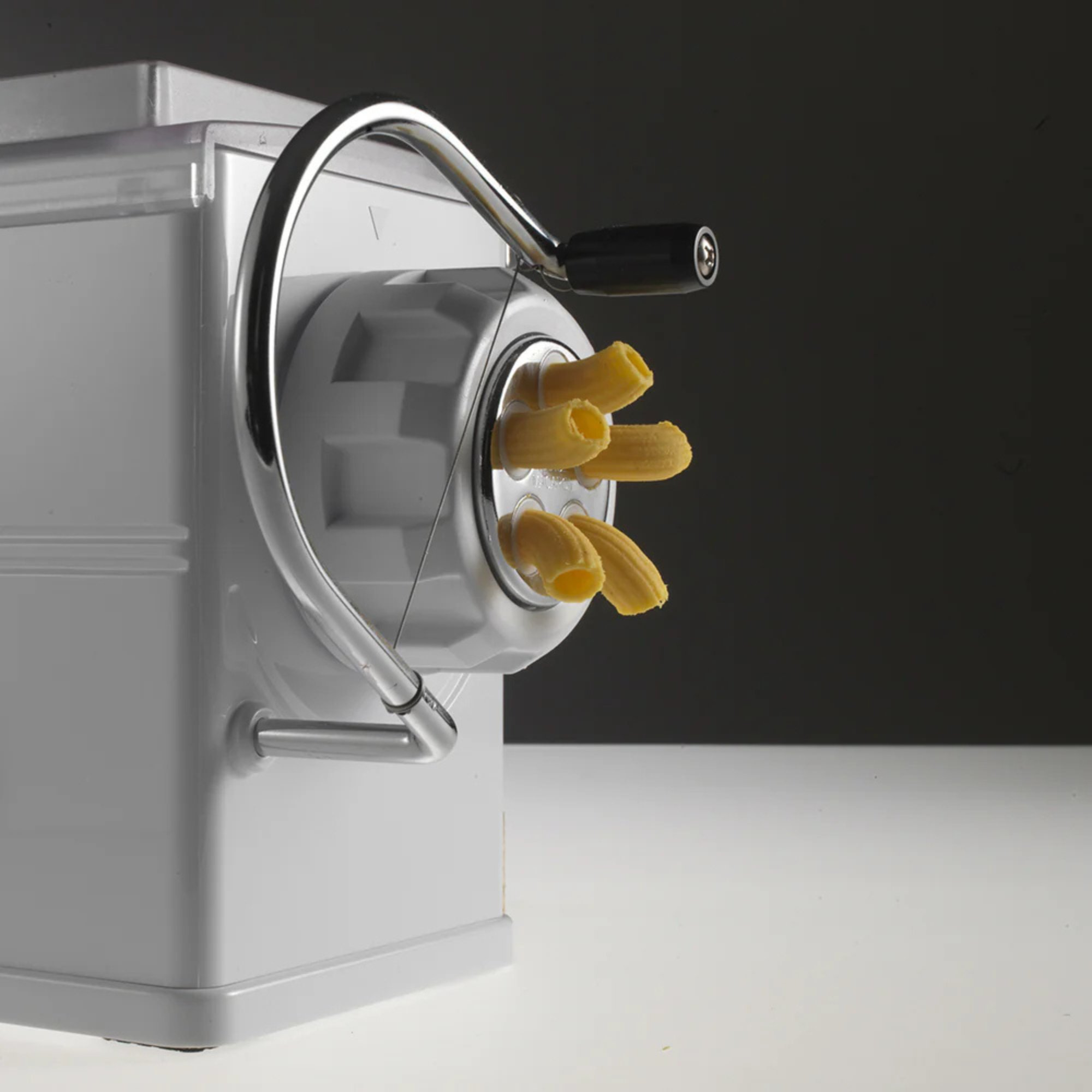 Eine weiße Nudelmaschine mit einem silbernen Griff und frisch extrudierten Rigatoni-Nudeln.