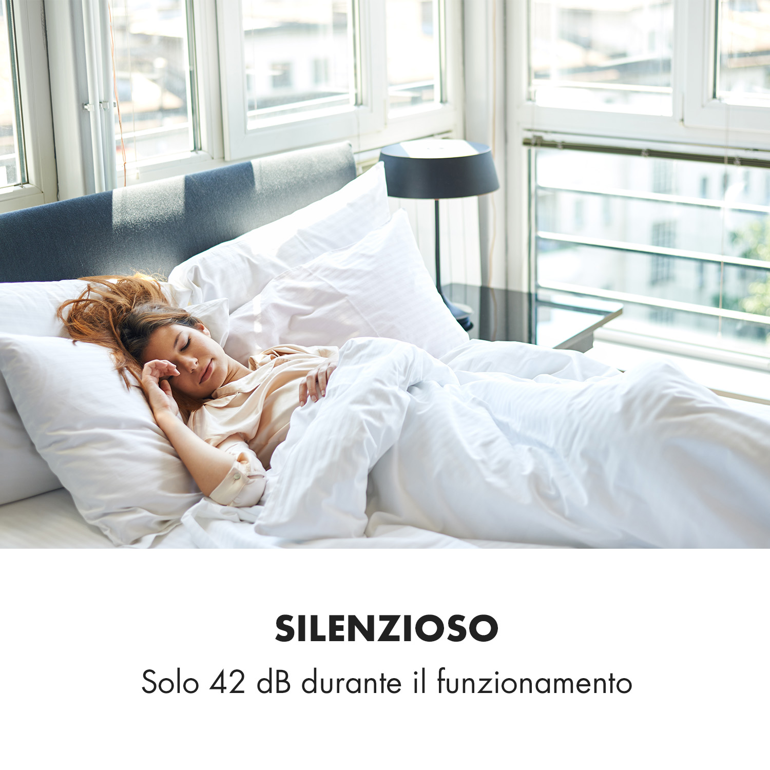 Una mujer duerme en una cama cubierta con sábanas blancas, frente a ventanas.