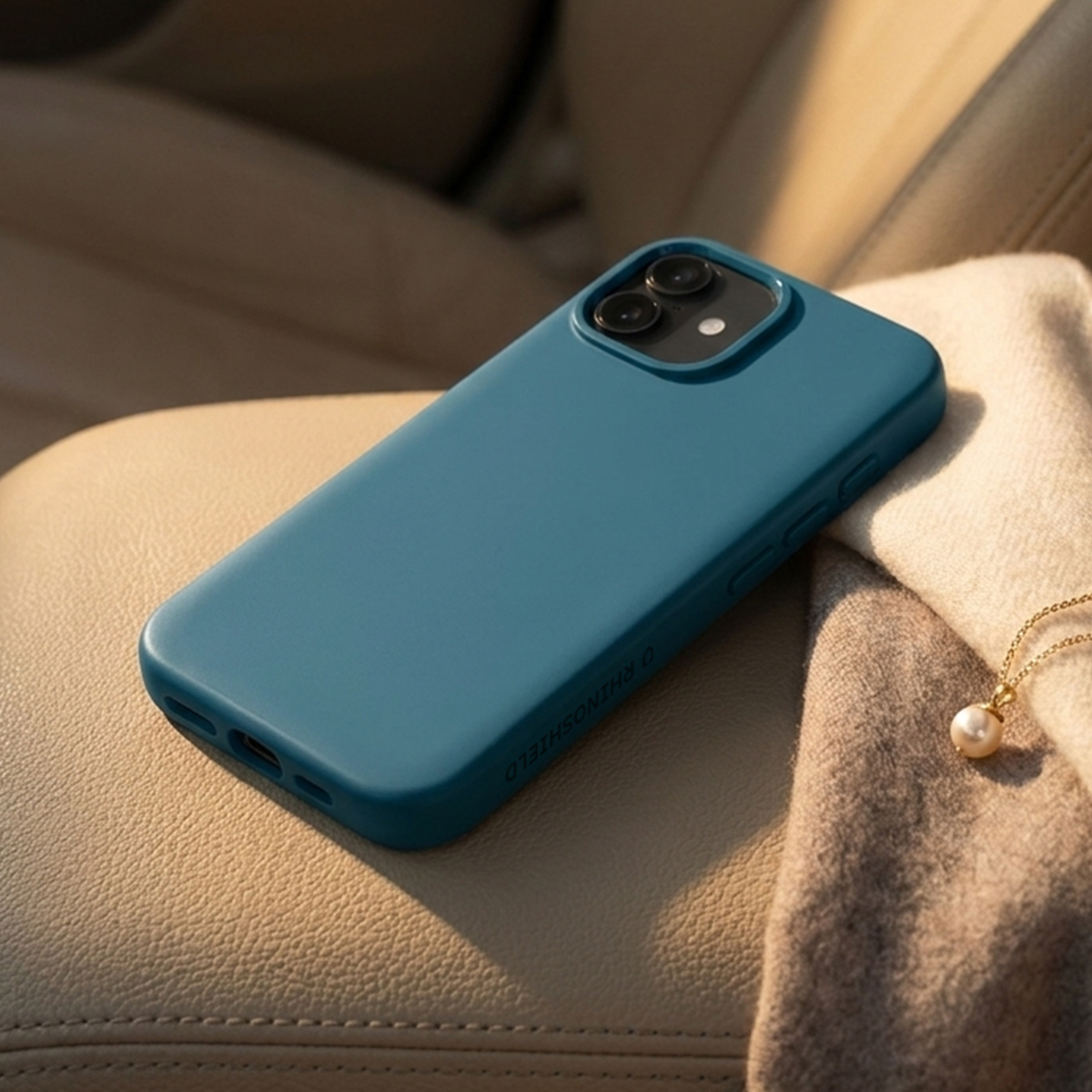 Una custodia blu petrolio per iPhone poggia su una sciarpa color crema, con una collana di perle nelle vicinanze.