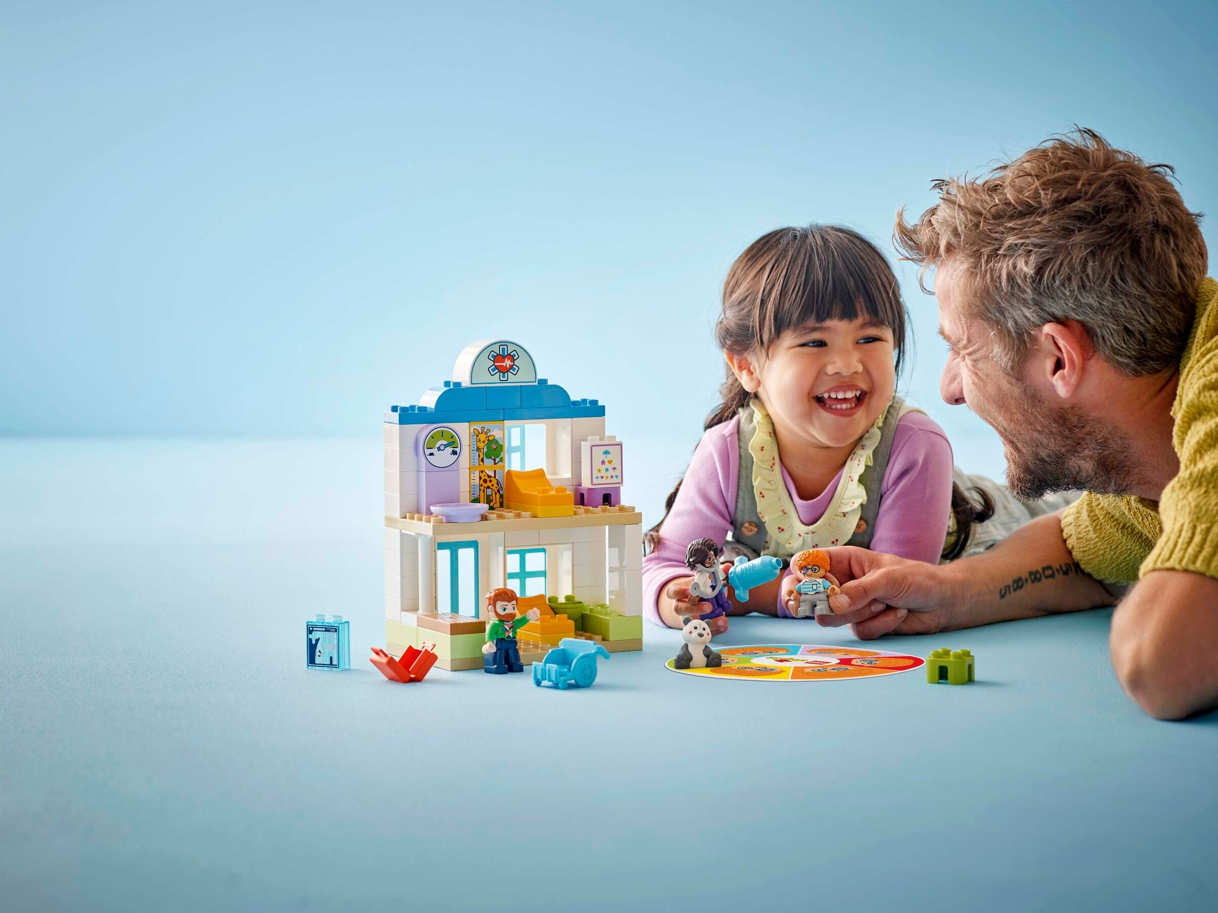 Ein Mann und ein Mädchen spielen mit einem Legohaus. Ein Panda sitzt auf einem Spielbrett.