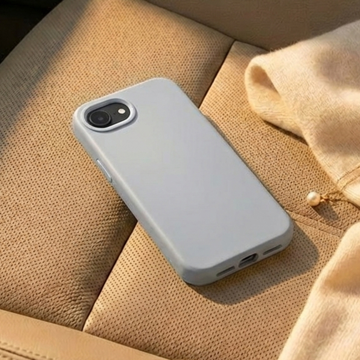 Una cover grigio chiaro per telefono poggia su un sedile auto in pelle traforata color cuoio. Accanto una sciarpa beige.