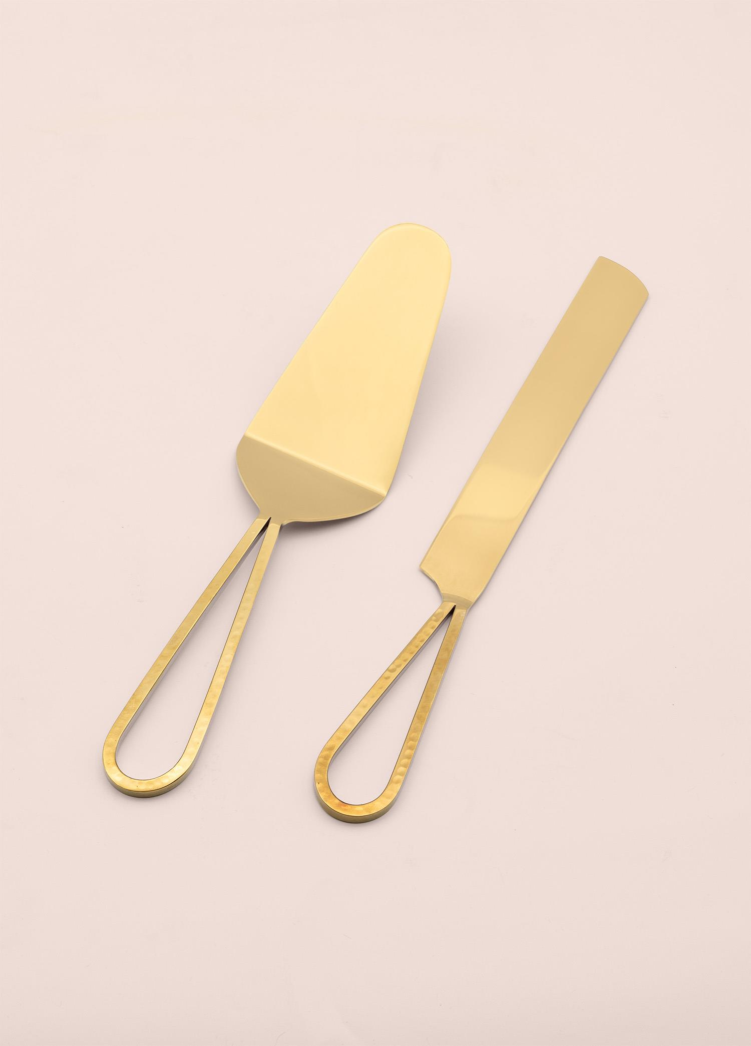 İki altın rengi pasta servis bıçağı açık pembe zeminde duruyor.