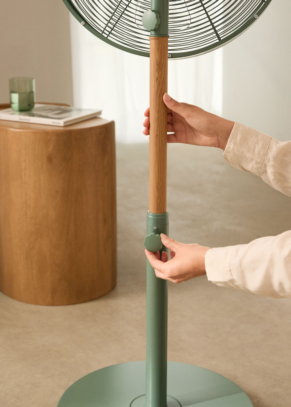 Manos ajustan la altura del ventilador con una perilla verde. Acento de madera en el soporte.