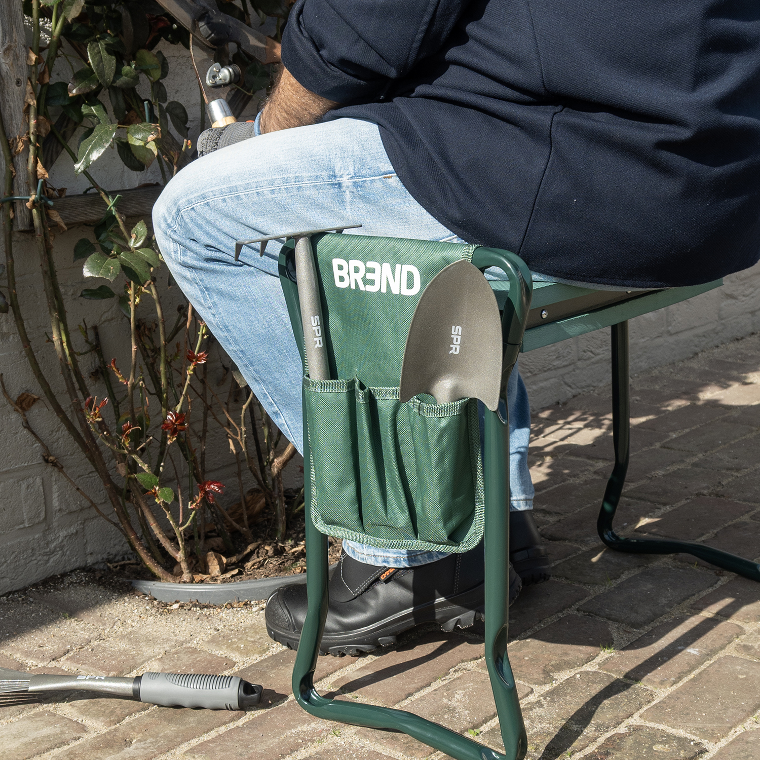 Homme assis sur un banc de jardin vert avec pochette à outils; outils de jardinage visibles.