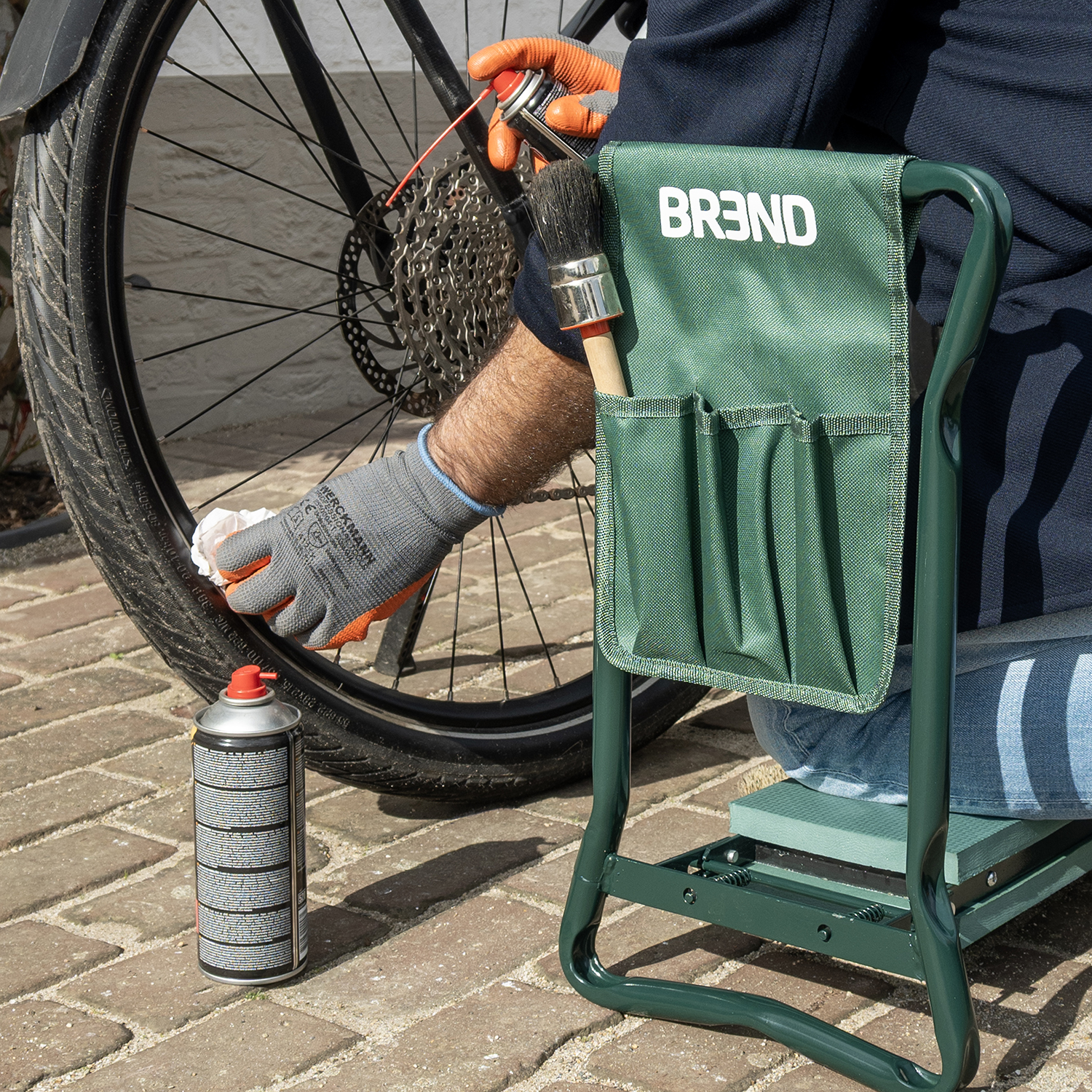 Personne gantée nettoie chaîne de vélo avec spray et chiffon, assise sur tabouret vert.