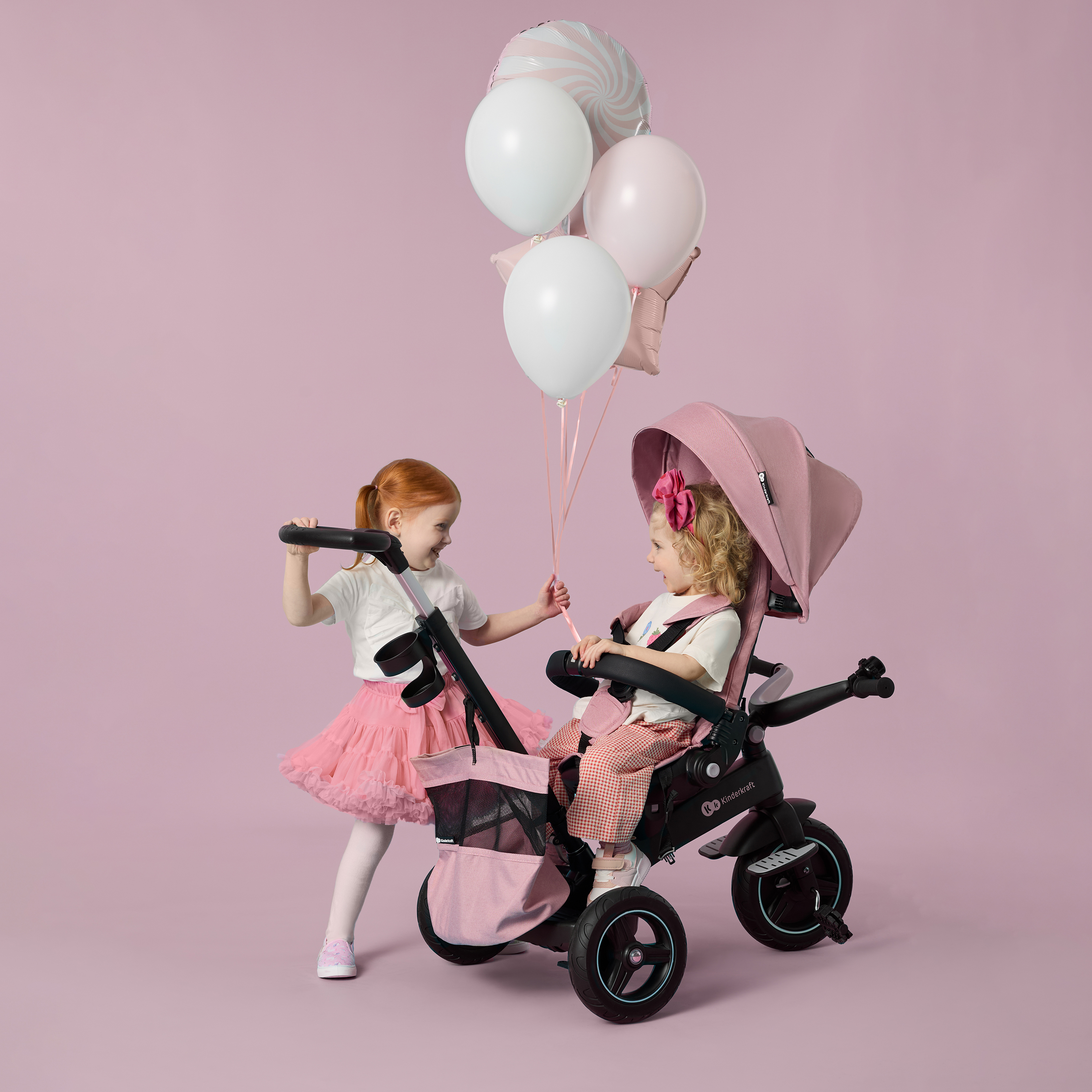 Zwei Mädchen mit Ballons und einem rosafarbenen Dreirad vor einem rosa Hintergrund.