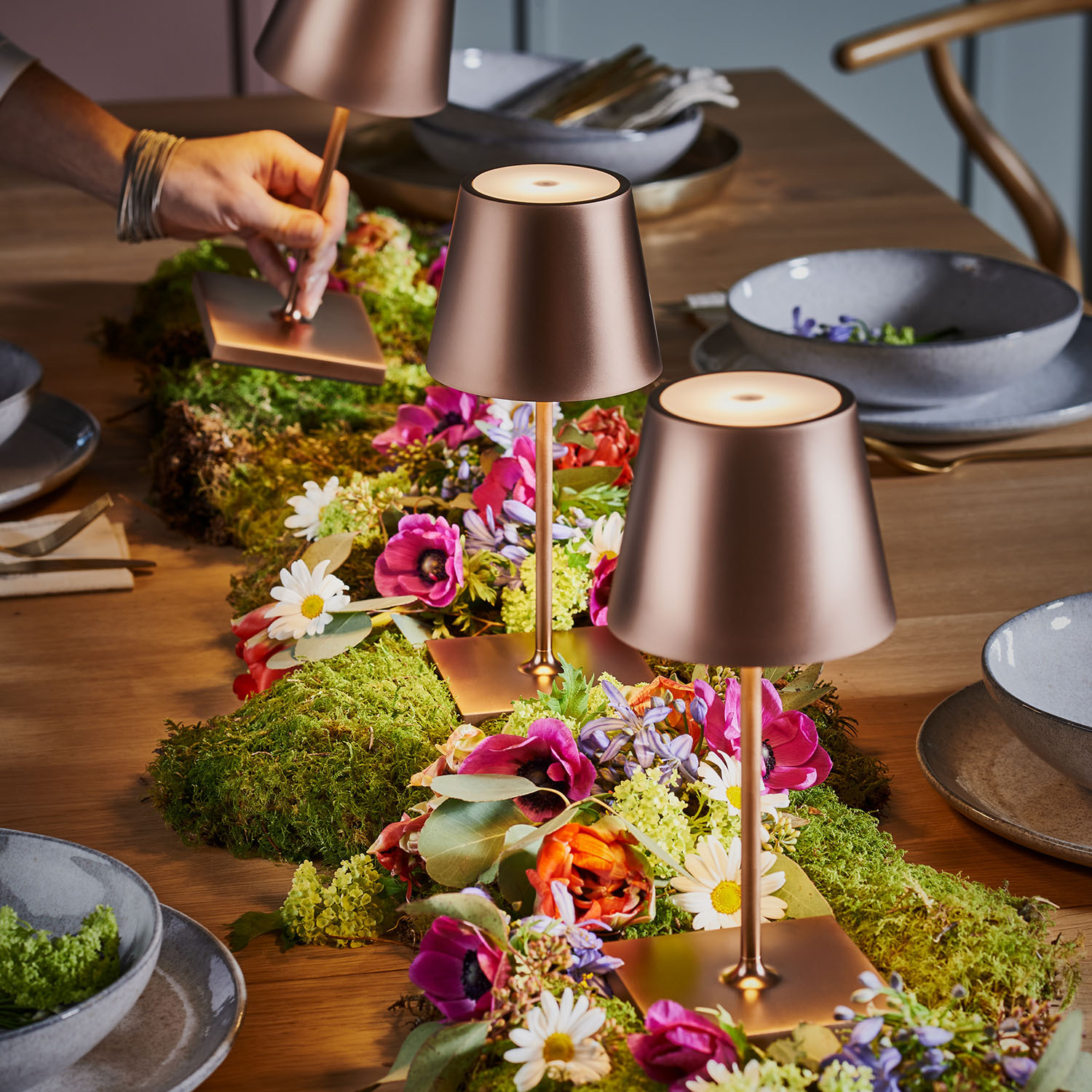 Ein Holztisch mit Blumen, Moos und zwei kupferfarbenen Lampen.